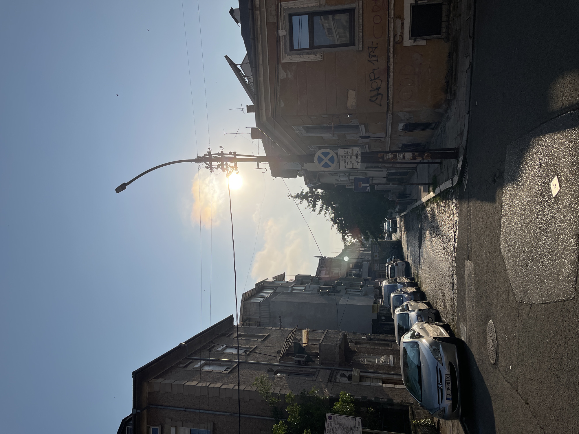 This image depicts a sunlit street scene in the city of Burgas, Bulgaria, likely during early morning or late afternoon, as indicated by the low sun angle and long shadows. The location is confirmed by the Bulgarian license plate "A 3598 HK" on the silver Peugeot 307 in the foreground (where 'A' signifies Burgas region) and the Cyrillic text on a street sign, which translates to a "Zone controlled by heavy vehicles type 'Spider' (i.e., tow truck)."
The sky is a clear light blue with some scattered white clouds, particularly visible around the bright sun, which is positioned in the upper-middle right, creating a visible lens flare and cast over the scene. Several small, dark birds are visible in the upper sky.
On the left side of the street, a row of multi-story residential buildings with various balconies and visible exterior conduits line the road. The closest building has a textured grey facade. Below these buildings, multiple cars are parked parallel to the curb, starting with the silver Peugeot 307 in the immediate foreground, followed by several other vehicles receding down the street. The sidewalk along this side is partially visible, with some greenery.
The street itself begins with an asphalt surface in the foreground, featuring a manhole cover near the center, and transitions to a cobblestone surface further into the mid-ground, curving slightly to the left as it recedes. Patches of the street appear reflective due to direct sunlight.
On the right side, an older, two-story building with a faded orange-yellow facade and significant peeling paint occupies the corner. Graffiti, including "GHOP 197. COD3," is visible on its lower walls. One window on this building reflects an architectural structure across the street. A tall, dark street light pole with a curved arm extends over the street from this side. Directly behind it, a utility pole with multiple cross arms and numerous overhead wires is present, with wires traversing across the sky. Several blue and white circular street signs are affixed to this pole, one of which identifies the "tow away zone." Further down the right side, a tall conifer tree is visible, along with other buildings and a sidewalk where a solitary figure is walking away from the viewer in the distance.
The overall impression is of an urban residential street with a mix of older architecture and modern vehicles, bathed in strong, directional sunlight.