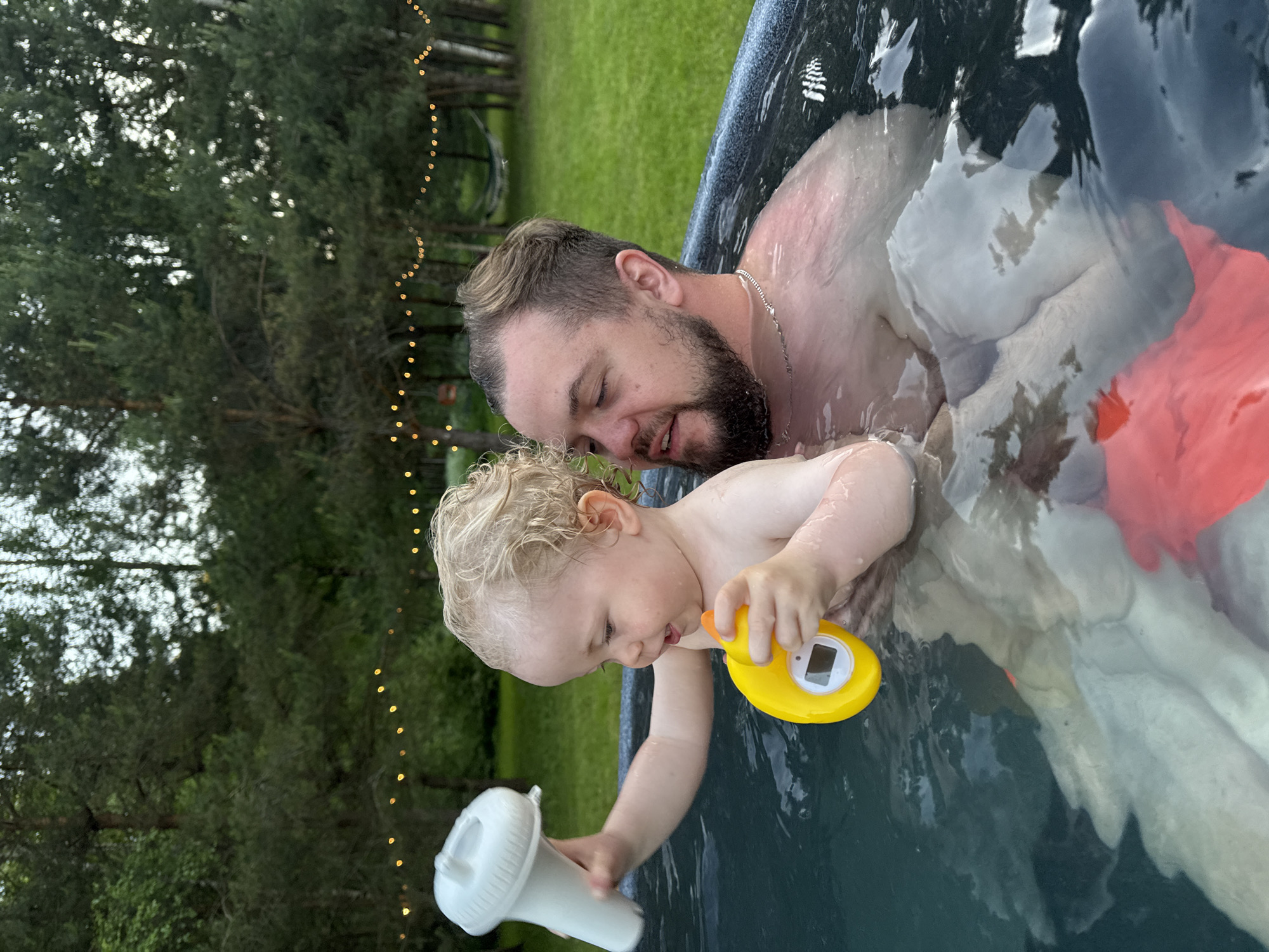 An adult male and a toddler are partially submerged in a dark-colored outdoor tub or small pool, located in an outdoor setting in Копнино, Russia. The adult male, positioned on the right, has short brown hair with lighter highlights, a beard, and a silver chain around his neck. He is looking down and to his left, towards the child. The toddler, positioned centrally on the left, has curly blonde hair and is holding two bath toys: a white cylindrical object with a textured cap in their raised left hand, and a yellow duck-shaped object with a small digital display in their right hand. The child's mouth is slightly open, and they are looking down at the yellow toy. The water level covers the chests of both individuals. In the background, numerous green trees, possibly conifers, form a dense canopy. A string of warm-toned decorative lights is strung horizontally between some of the trees. A white hammock is visible hanging between two trees in the upper right background. A patch of green grass is visible behind the tub.