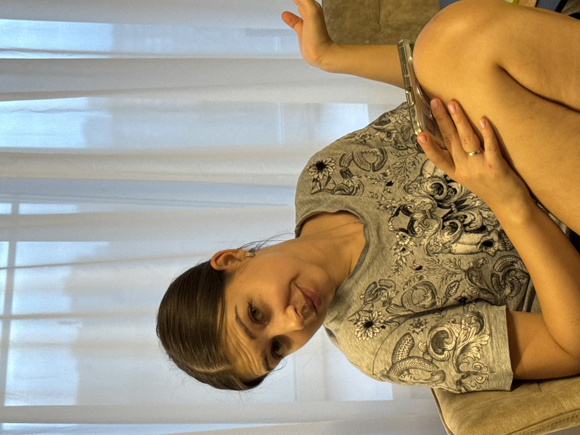 A woman with brown hair is sitting indoors, holding a smartphone. She is wearing a gray T-shirt with a floral and snake pattern. The background features a white window blind. No information on the city of Krasnoyarsk, Russia is available in this image.
