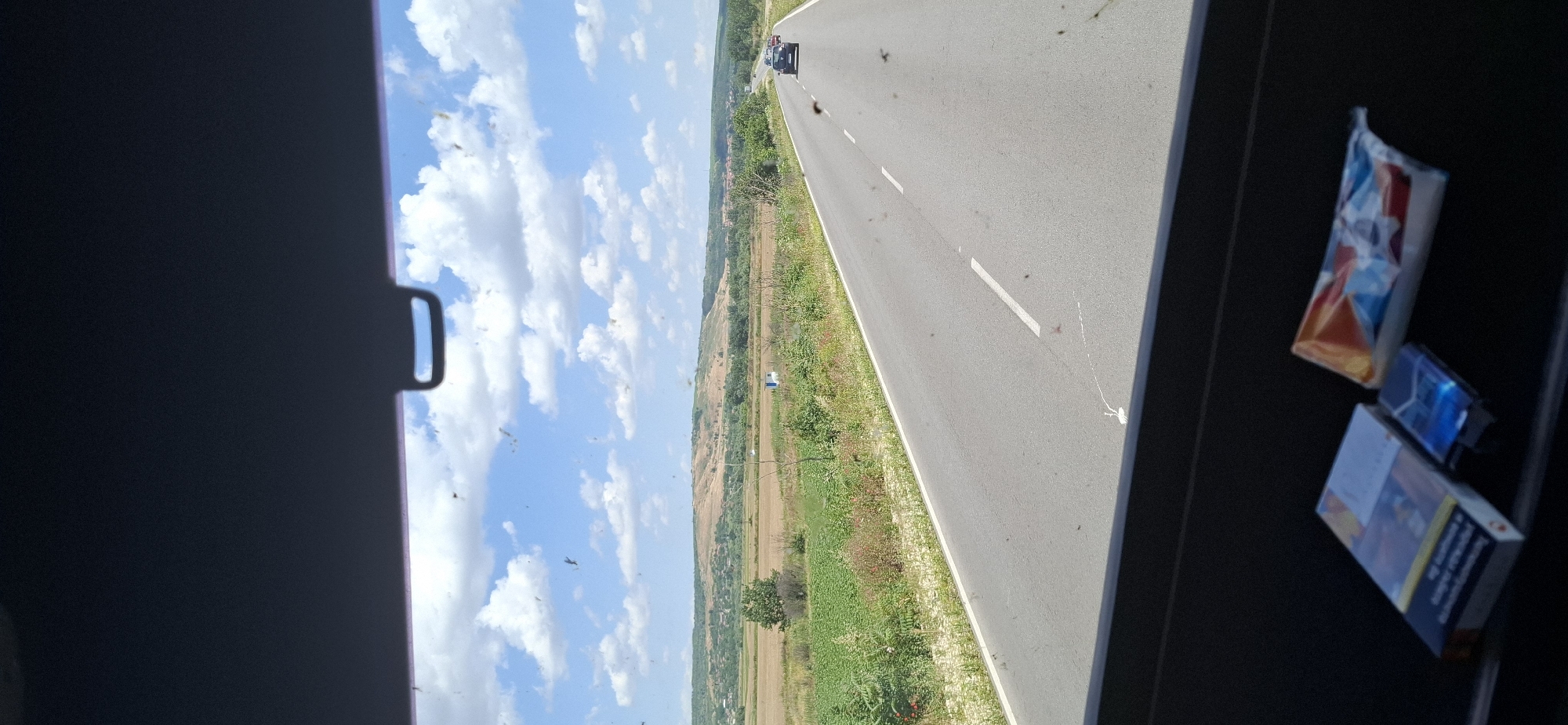 The media file depicts a view from inside a vehicle, looking out at a rural road in Opanets, Bulgaria. The scene is captured in a vertical orientation, suggesting the photo was taken while the vehicle was in motion or the camera was held sideways.
The main subjects are the road and the surrounding landscape. The road is asphalt with white dashed lines marking the lanes, stretching into the distance. A few vehicles are visible on the road, including a black car ahead and what appears to be a white van further back. The landscape on either side of the road consists of fields, some with dry, golden crops and others with green vegetation. Trees and bushes line the roadside and dot the fields. In the distance, rolling hills covered in greenery are visible. The sky is a bright blue with scattered white, fluffy clouds.
The activity taking place is travel along the road. The presence of vehicles and the linear road suggest a journey.
Notable details include the time of day, which appears to be daytime due to the bright sunlight and clear sky. The weather seems to be fair and sunny. The location cues, such as the rural setting and the text on the cigarette packs, confirm the location as Opanets, Bulgaria. There are also several small, dark specks visible on the windshield or window, possibly insects or dirt.
On the right side of the frame, on a dark surface, are two packs of cigarettes. One is a lighter colored pack with a colorful abstract design, and the other is a darker pack with warning labels and text in a language that appears to be Cyrillic, consistent with Bulgarian. Some of the visible text on the darker cigarette pack includes warning messages and possibly brand names, but the text is too blurry to be fully legible. The other pack has a predominantly blue, red, and orange geometric pattern.