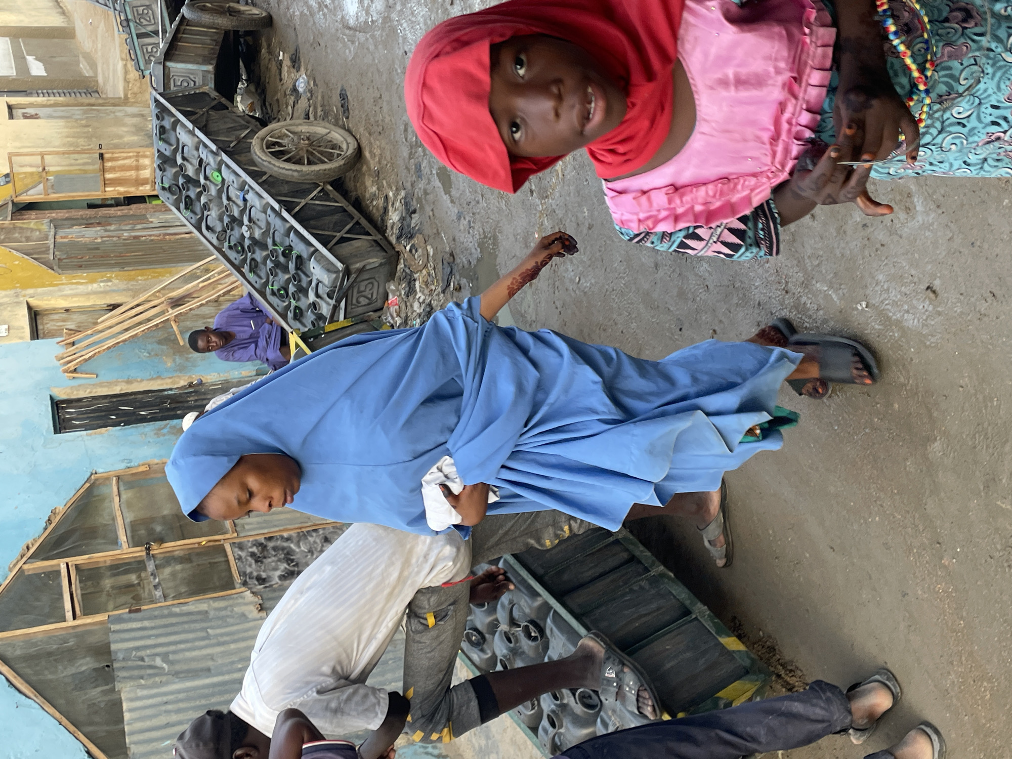 In an outdoor urban setting, possibly a street, individuals are situated amidst objects suggesting commercial activity. A young girl wearing a red head covering and a pink patterned dress looks directly at the camera. Nearby stands a person dressed in a blue hijab and long blue gown. There are two men; one wearing a white long-sleeved shirt and light colored pants, another dressed in a purple long-sleeved shirt.
The area is congested with commercial objects including carts and bottle crates.