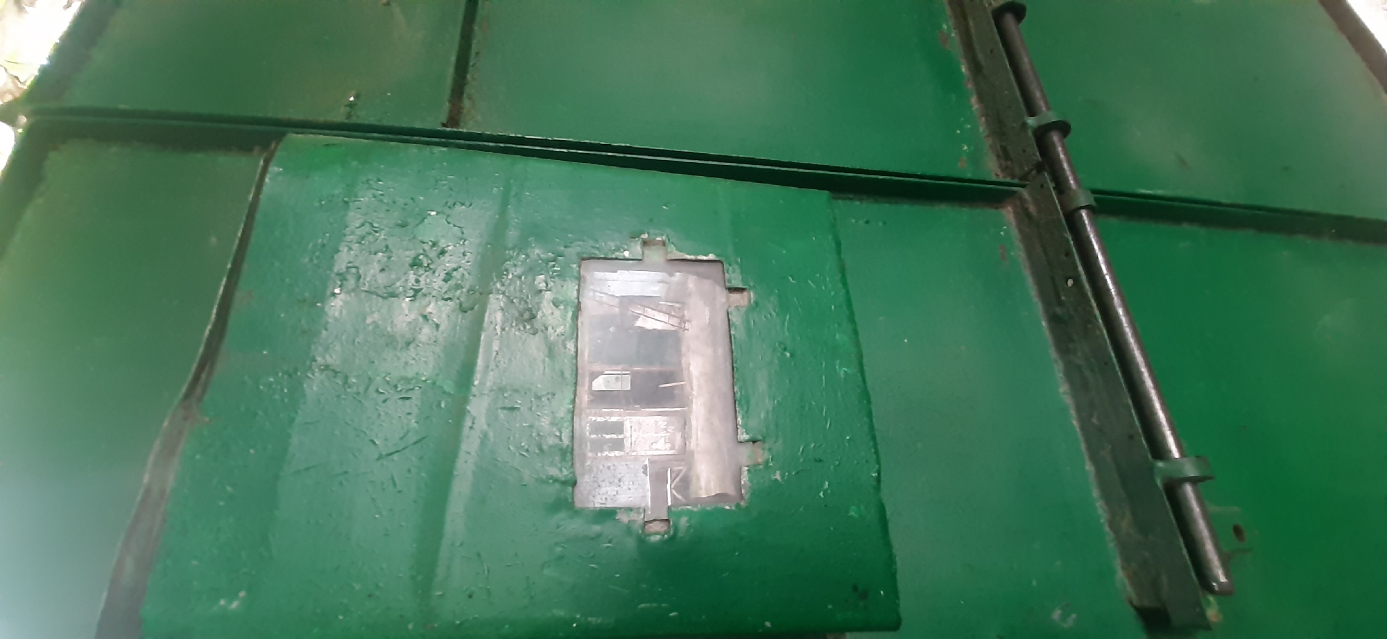 This image shows a close-up view of a large, weathered green metal surface, possibly a door or a panel of a structure. The surface is segmented by dark lines, indicating it is composed of multiple pieces. There is a small, rectangular window or opening in the center, made of glass or clear plastic, which appears to be dirty and smudged. Inside the opening, there are some indistinct, layered objects or reflections.
Along the right side of the image, there is a vertical metal rod or pipe secured by brackets to the green surface. This rod appears to be part of a locking or fastening mechanism. The green paint on the metal is chipped and worn in several places, revealing darker material underneath and suggesting age and exposure to the elements. The overall impression is one of industrial utility or a protective barrier.
The setting is not explicitly shown, but the object itself could be part of a ship, a train car, a bunker, or an industrial building. The lighting suggests it is daytime, with no specific indication of weather conditions. The image does not contain any visible text. The location context provided is Yessentukskaya, Russia, which could imply this is an object within that geographical area.