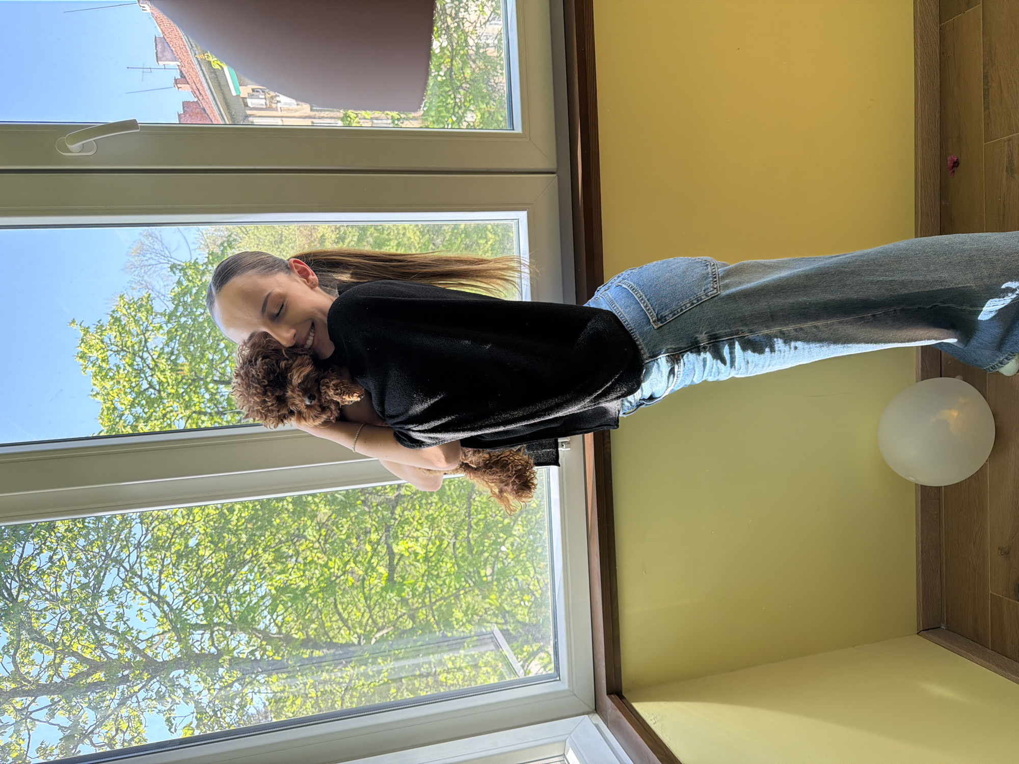 A woman in a black sweater and blue jeans is holding a brown poodle near a window. The window looks out onto a scene of green trees and a blue sky. The interior walls are painted a light yellow. There is a white balloon in the corner of the room. The location is likely Sofia, Bulgaria, based on the building architecture seen through the window.