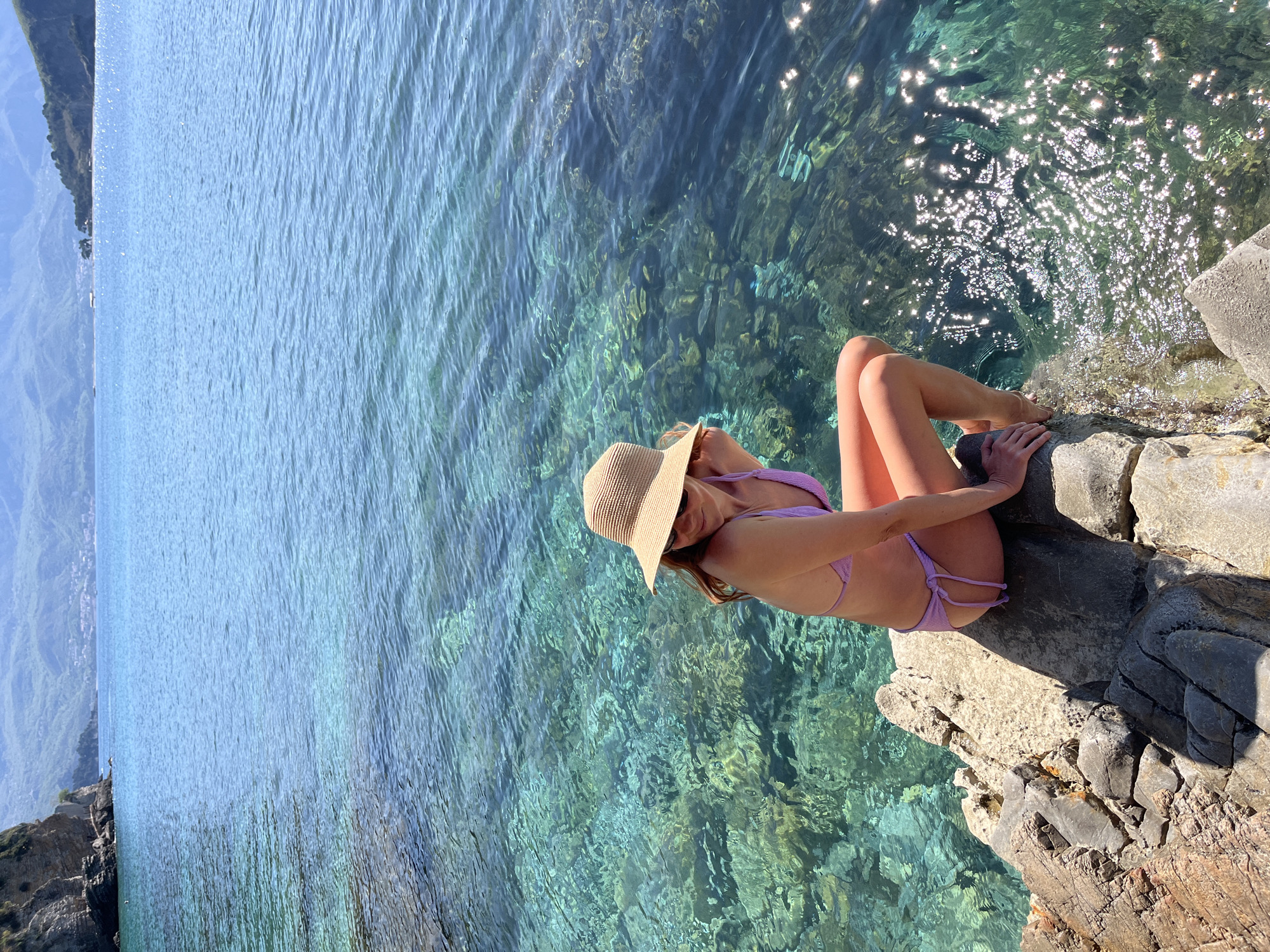 A sunlit, elevated shot of a woman in a lavender bikini and a straw hat sitting on a rocky outcropping, with the sparkling turquoise waters of the Adriatic Sea below. The outcropping appears to be on a cliff or a rocky edge, with sunlight reflecting off the water. The sky is visible in the distance. The scene suggests a coastal location, potentially near Budva, Montenegro, given the characteristics of the water and coastline.