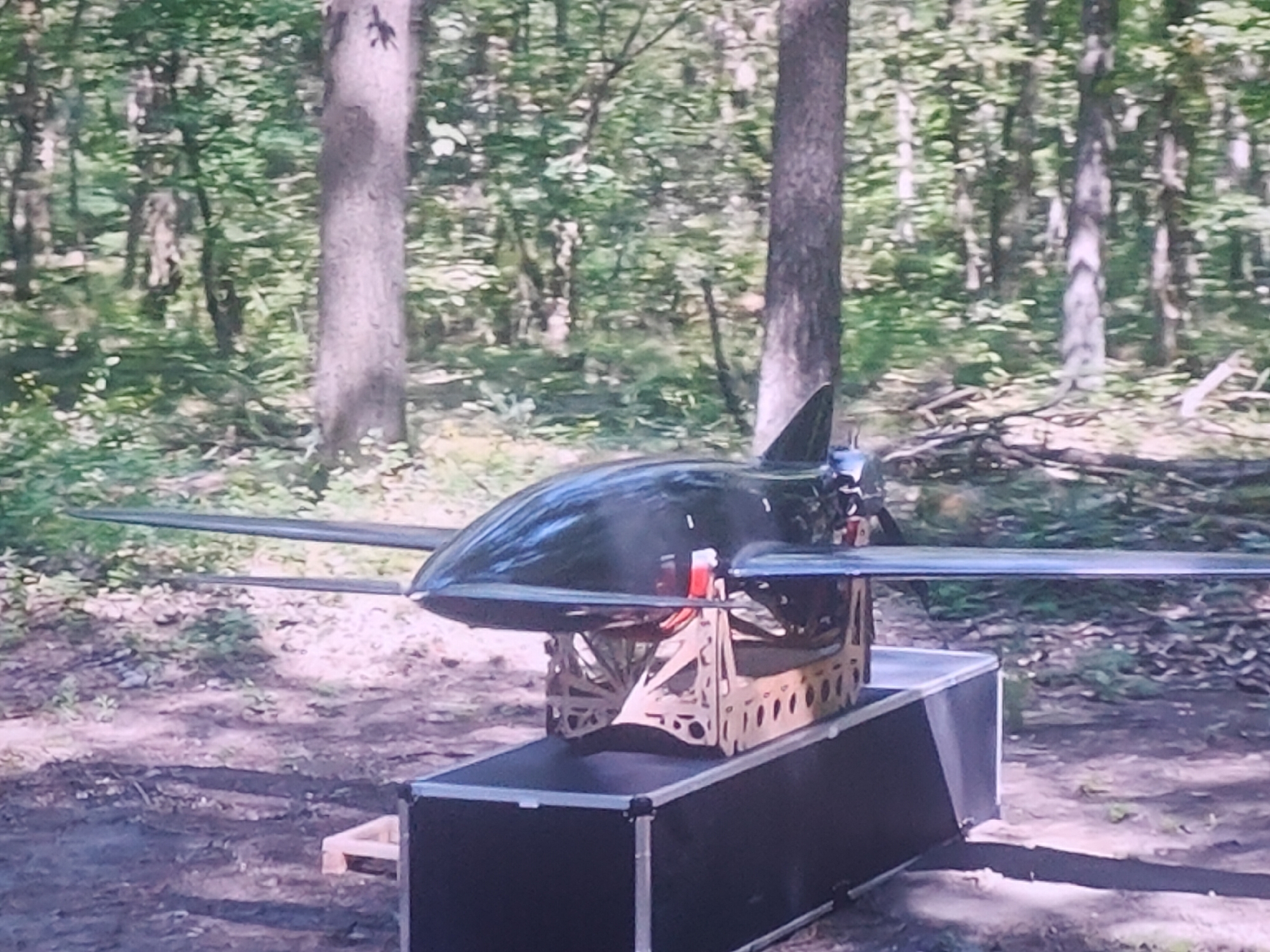 A drone with swept wings and a pointed nose is mounted on a complex wooden cradle which is itself resting on a black equipment case. The drone is dark, possibly black or very dark grey, and appears to be made of a smooth, reflective material. The cradle has an intricate, geometric design with numerous cutouts. The equipment case is rectangular with silver latches visible on its front. The scene is set in a wooded area with tall trees and dappled sunlight filtering through the leaves. The ground is covered with dirt, leaves, and small branches. The image quality is grainy and appears to be a photograph taken from a distance or with a low-resolution camera. There are no people visible in the image. The location is stated as Molenbeek-Saint-Jean - Sint-Jans-Molenbeek, Belgium, but this is not visually evident from the image itself.