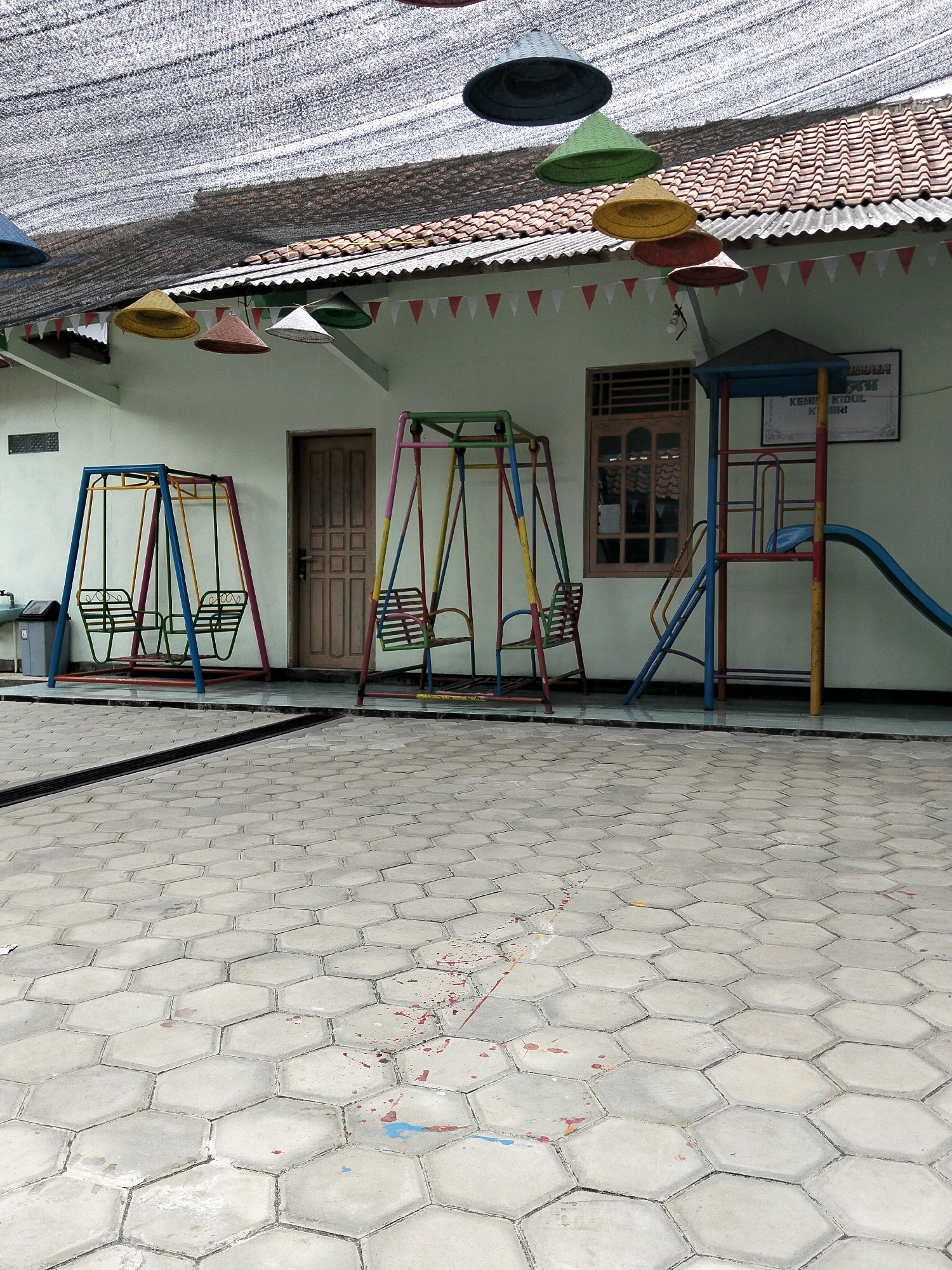 The image depicts a playground in Krajan Lor, Indonesia, with a building in the background. The playground features two colorful metal swingsets, one with two seats and the other with one seat. Adjacent to the swingsets is a small play structure with a blue slide. The ground is paved with hexagonal gray tiles, with some red and blue paint splatters visible on the tiles. Above the playground, a black mesh shade is stretched across, supported by a tiled roof. Several colorful, conical hats hang suspended from the shade. A wooden door and a window are visible on the building's exterior. A trash can is located to the left of the swingsets. A sign is posted on the wall of the building.