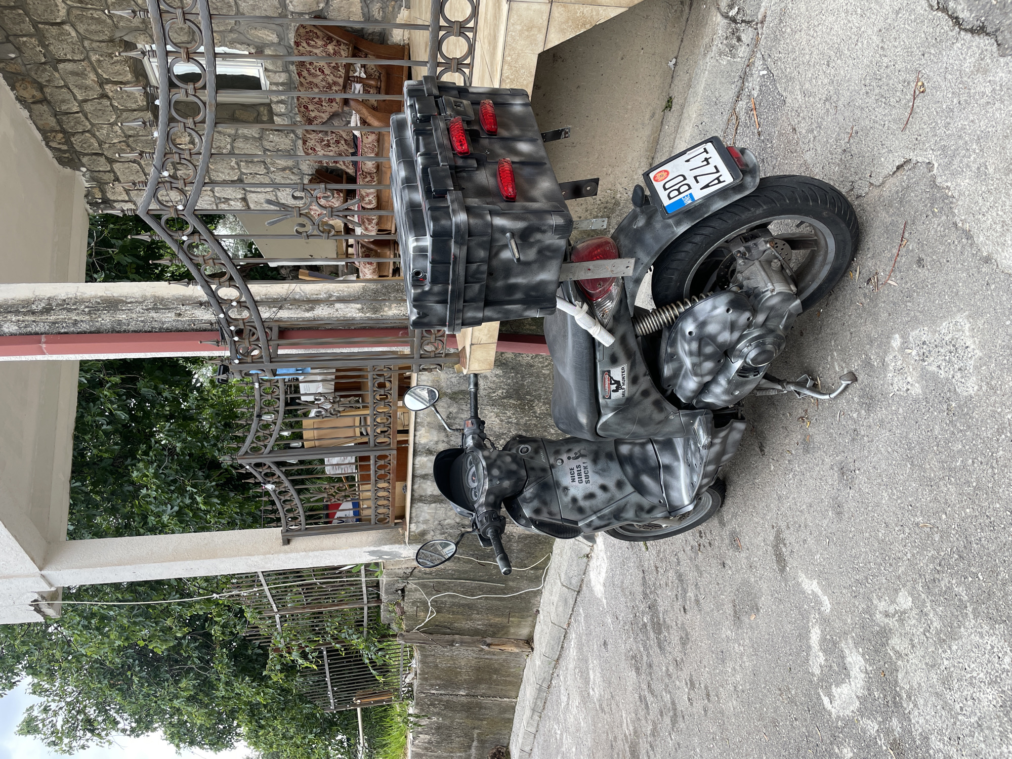 A grey and black camouflage-painted scooter is parked on an asphalt road in Rafailovići, Montenegro, next to a building with a stone and concrete exterior. The scooter has a large, boxy storage compartment mounted on the back and a license plate that reads "BD A241". The background includes a metal fence, foliage, and a partial view of a building.