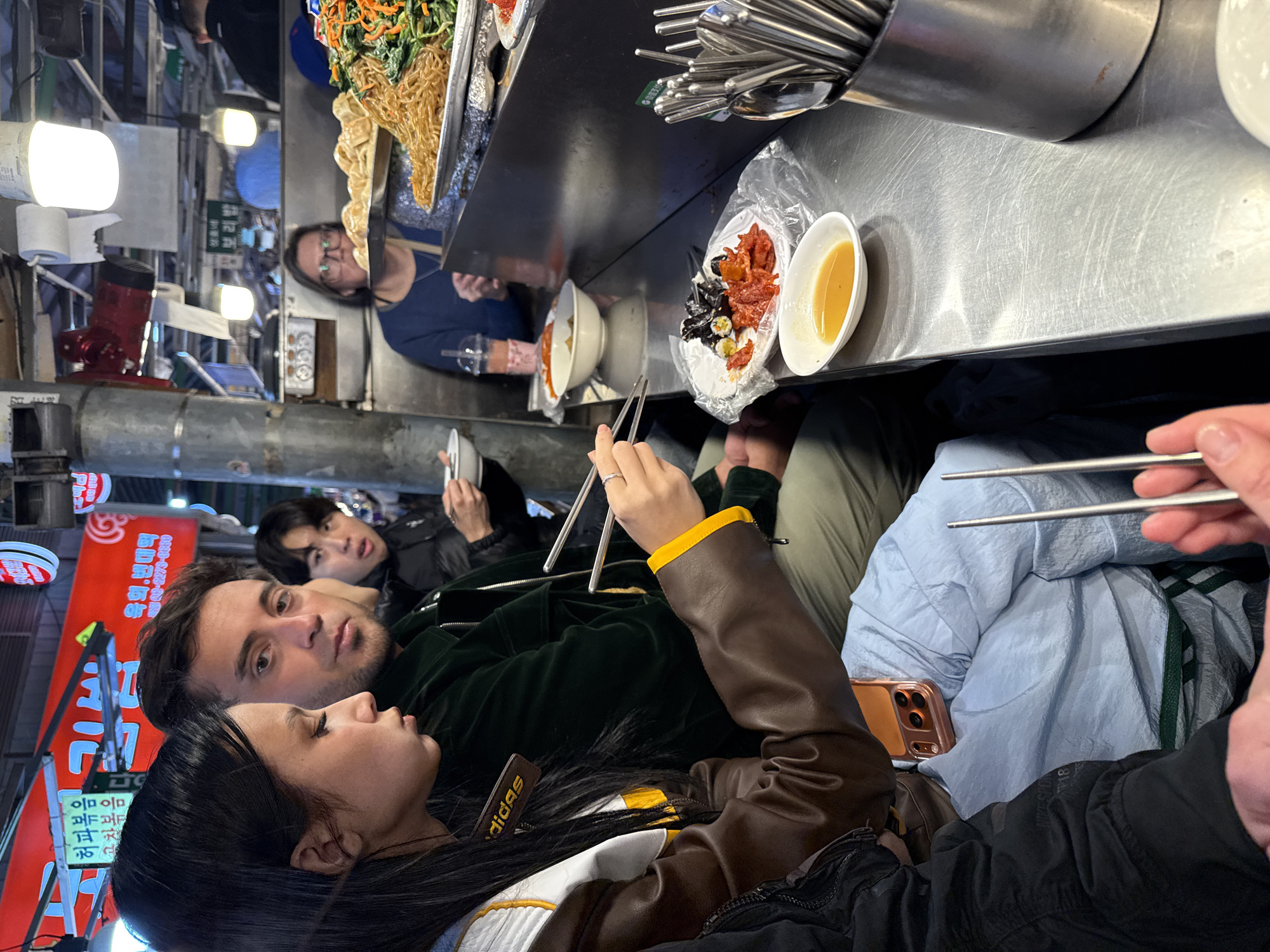 The image depicts an active street food stall located in a bustling market area of Seoul, South Korea. This is identified by the Korean script on overhead red awnings (e.g., "해피볶음", "김밥", "보리밥") and the characteristic presentation of Korean street food.
At least five individuals are visible within the frame, primarily focused on eating. In the foreground, a female with long dark hair, wearing a brown and white leather jacket with yellow accents (an "Adidas" tag is visible near her neck), is holding metal chopsticks over a white plate containing kimbap (seaweed rice rolls) and a red stir-fried dish, possibly tteokbokki. This plate rests on a clear plastic wrap. A small oval bowl of yellow dipping sauce is positioned next to her plate on the polished stainless steel counter. An orange-cased smartphone is resting on a light blue garment draped below her.
Seated immediately to her right is a male with dark hair and a beard, wearing a dark green jacket, looking directly towards the viewer. Behind him, another male in a dark jacket is visible, actively eating from a white bowl with chopsticks.
Behind the counter, a female wearing glasses and a blue top is visible, positioned near the food display. Further back and partially obscured is another male wearing a dark cap.
The food display consists of large metal trays filled with various Korean dishes, including japchae (glass noodles with vegetables), mandu (dumplings), and what appears to be an assortment of banchan (side dishes). A stainless steel container filled with metal chopsticks and spoons is prominent on the right side of the counter. A translucent plastic cup containing a pink beverage with a straw is also visible.
The environment is brightly lit by overhead fluorescent fixtures. Structural elements include an exposed metal support pole. Rolls of white paper are affixed to the overhead structure. The overall scene suggests a casual, busy dining experience typical of a Korean market.