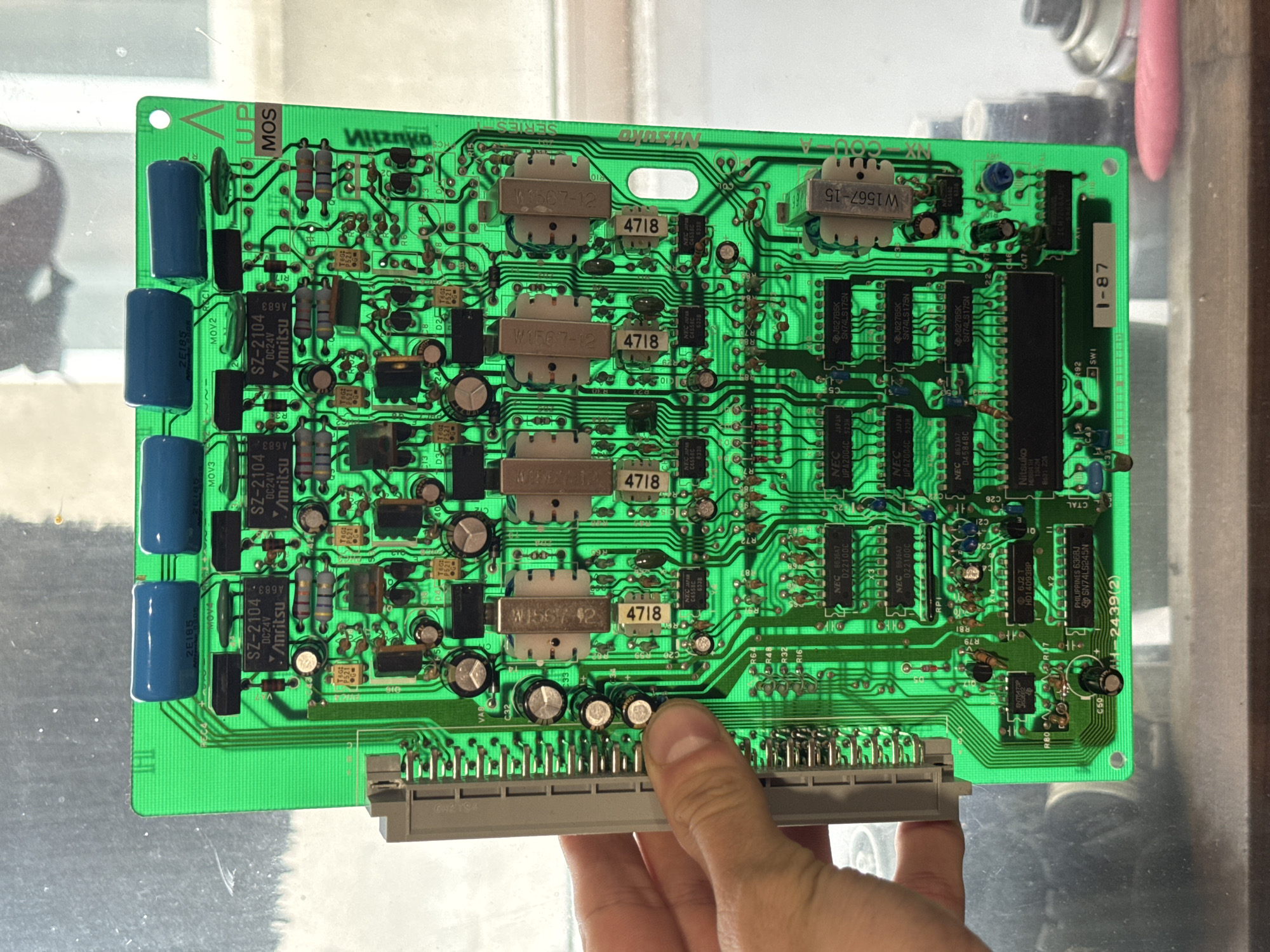 An overhead, close-up shot shows a green circuit board with numerous electronic components. The circuit board is illuminated from behind, making the components visible. Several blue, cylindrical capacitors are at the top. Below them are rows of black, rectangular integrated circuits and smaller, silver components. Larger, cream-colored components with the text "W1567-12" are present in the middle. Resistors, diodes, and numerous smaller capacitors are also visible. The circuit board has some printed text, including "Nitsuko" and "1-87". A grey connector is attached to the left edge of the board. A person's hand is visible on the left, holding the circuit board. The background appears to be a window with an out-of-focus view of a cluttered area, possibly a workshop or storage space. The scene is indoors.
