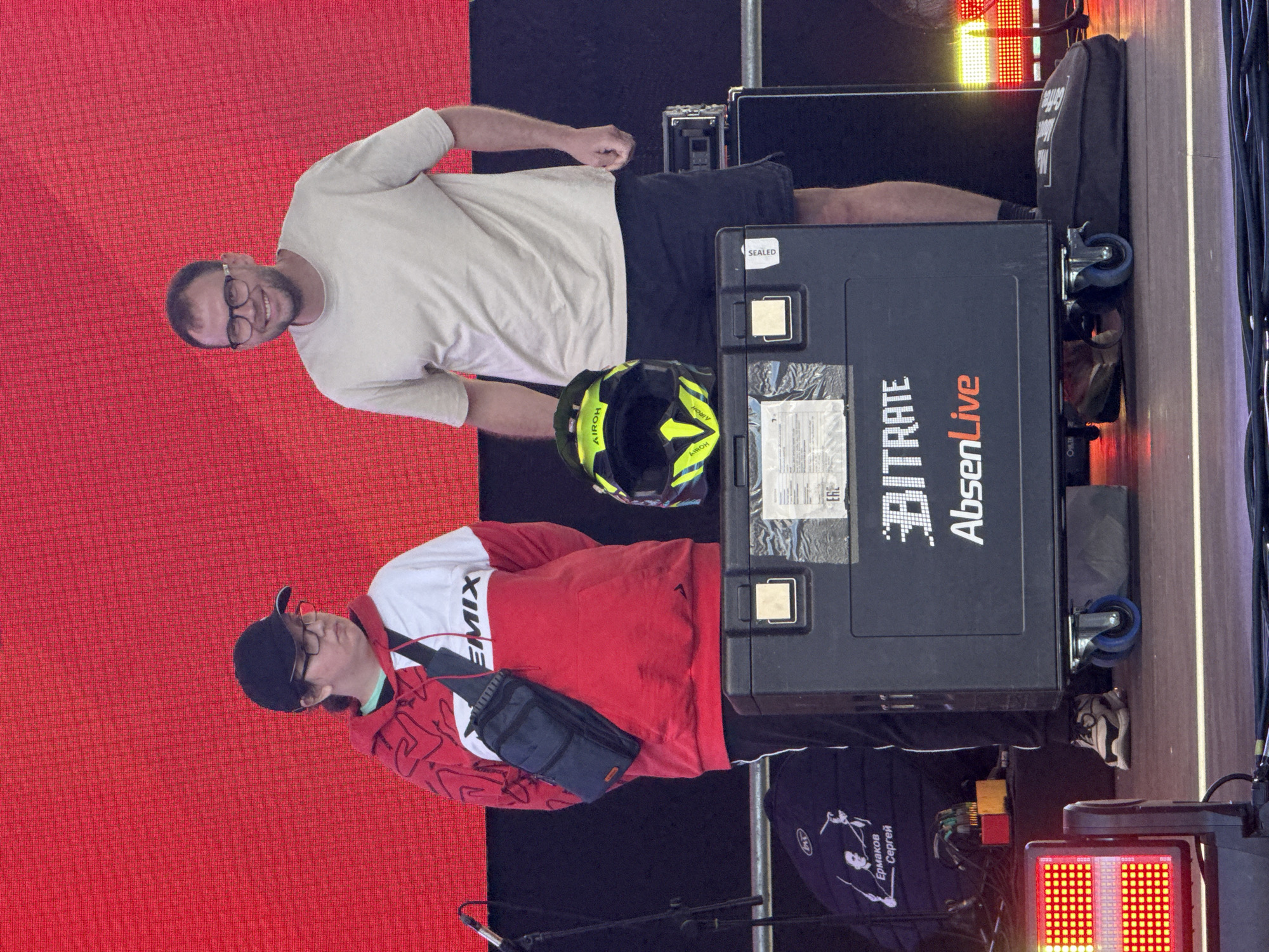 The image captures two individuals standing on a stage in front of a large red LED screen. On the left, a person wearing a black baseball cap, glasses, and a red and white hoodie with a black sling bag over their shoulder is looking slightly to their right. On the right, a man wearing glasses and a light-colored t-shirt and dark shorts is smiling and looking forward. Between them is a black case on wheels, with a yellow and black helmet placed on top. The case has the text "BITRATE AbsenLive" printed on it. To the left of the case, a black bag with a graphic of two figures and Russian text "Ермаков Сергей" is visible, along with some smaller objects. On the right side of the stage, audio equipment and stage lighting are present. The overall environment suggests an outdoor event or concert setting, likely in Saint Petersburg, Russia, given the presence of Russian text on the bag.