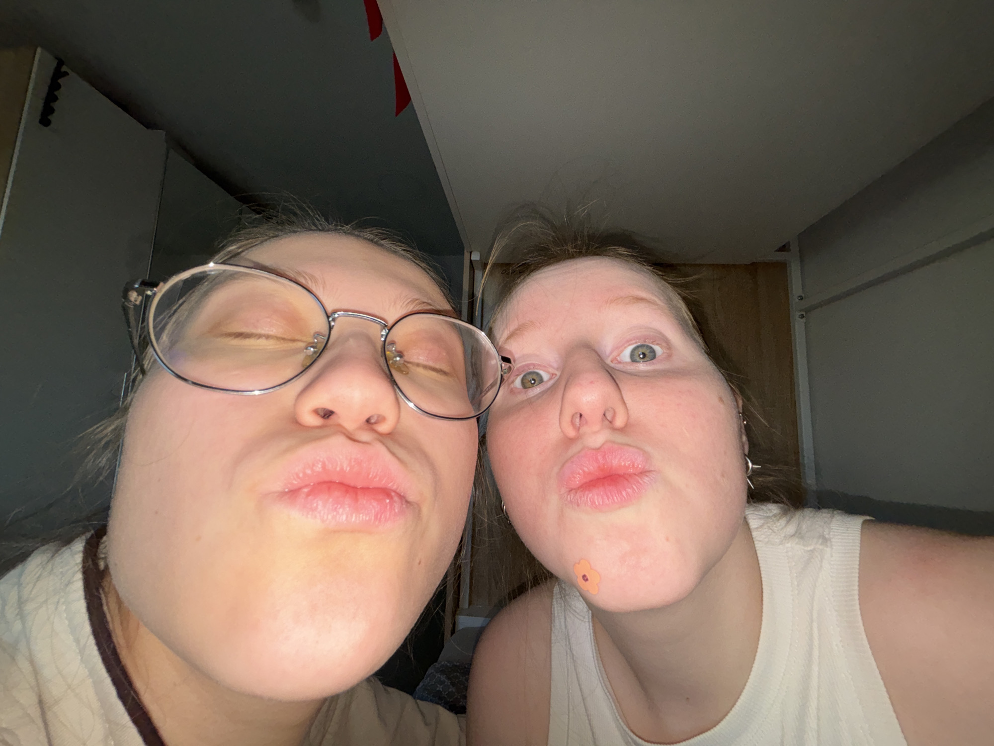 Two young women are posing for a selfie, puckering their lips towards the camera. The woman on the left is wearing glasses and has her eyes closed. The woman on the right has her eyes open and a flower-shaped sticker on her cheek. They are indoors, possibly in a bedroom, with a wardrobe visible in the background. The location is not evident in the image, and there is no information to determine if the photo was taken in Perm, Russia.