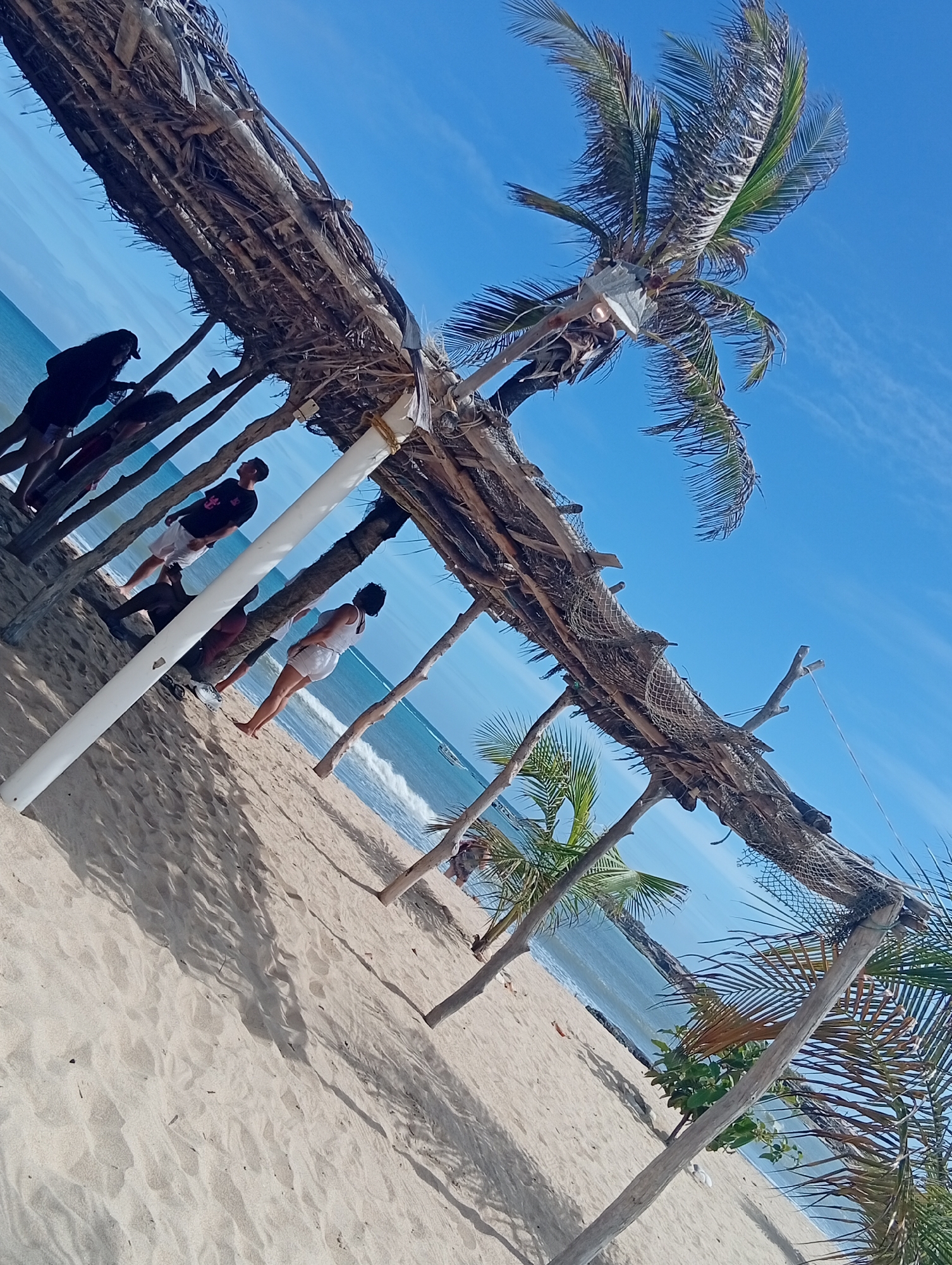 In El Tunal, Venezuela, a group of people are on a sandy beach beneath a rustic, open-air shelter constructed from palm fronds and driftwood. The shelter, supported by wooden poles and a white cylindrical post, provides shade from the bright sun. A tall palm tree with its fronds reaching towards the clear blue sky stands to the right of the shelter. The ocean is visible in the background with gentle waves lapping at the shore. Several individuals are standing or sitting within the shelter's confines, some looking out towards the sea, while others are engaged in conversation. The sandy ground is imprinted with shadows cast by the shelter and palm trees, suggesting a sunny day. The overall scene depicts a casual, relaxed atmosphere on a tropical beach.