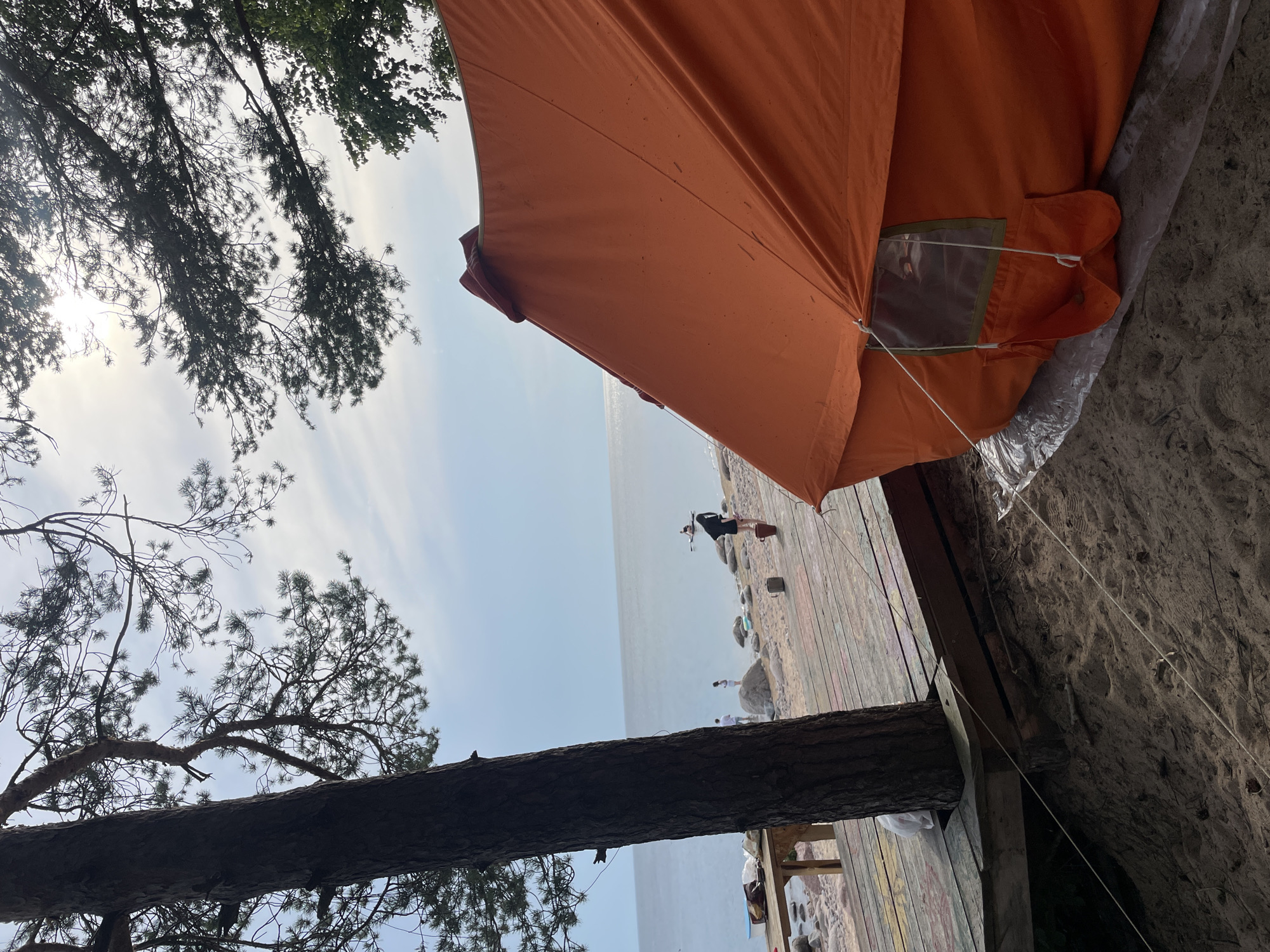 In Luzhki, Russia, an orange tent is pitched on a sandy shore near a wooden deck. The tent, positioned on the right side of the frame, is partially in shadow. A large tree trunk stands to the left, its branches extending overhead, partially obscuring a bright sky with scattered clouds. In the background, a body of water stretches to the horizon, with several people visible on the beach and in the water. One person stands near the water's edge, while another appears to be on a paddleboard further out. The wooden deck, decorated with colorful paint splatters, is located between the tree and the beach. A table with some items on it is also visible on the deck. The scene depicts a recreational area by the water on a day with moderate sunlight.