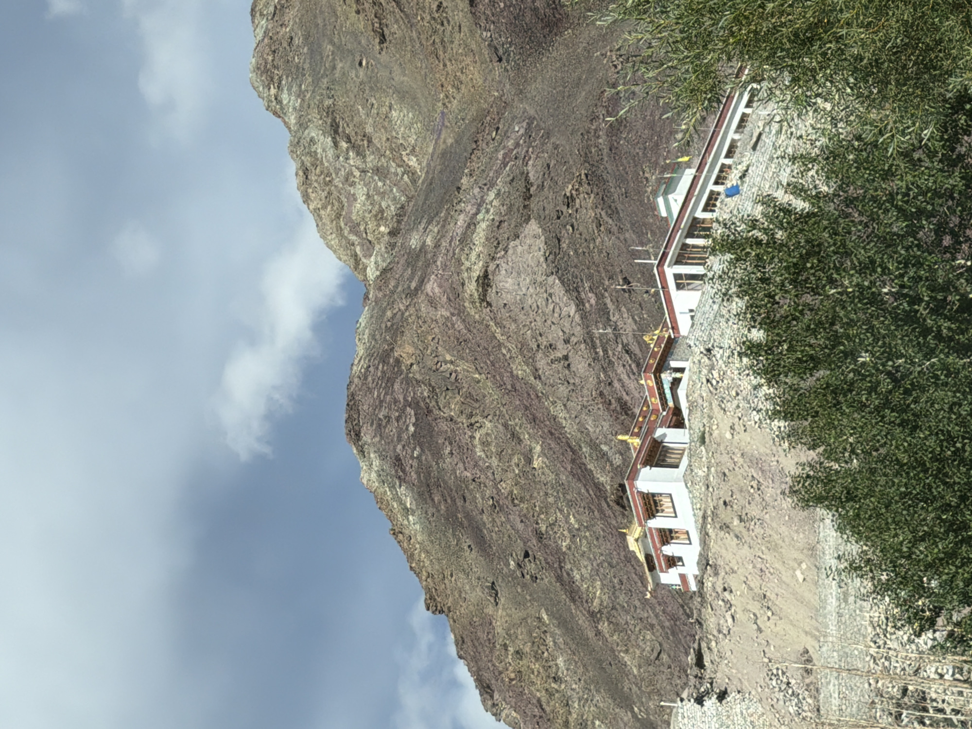 The image shows a multi-story building with white walls, red trim, and golden roofs situated on a rocky, barren mountainside. The building appears to be a monastery or temple, with prayer flags visible on the roof. The surrounding environment is dry and mountainous, with sparse vegetation in the foreground. The sky is partly cloudy. The building is located on the side of a steep, rocky mountain. The architecture is characteristic of Buddhist monasteries found in the Himalayan region.
