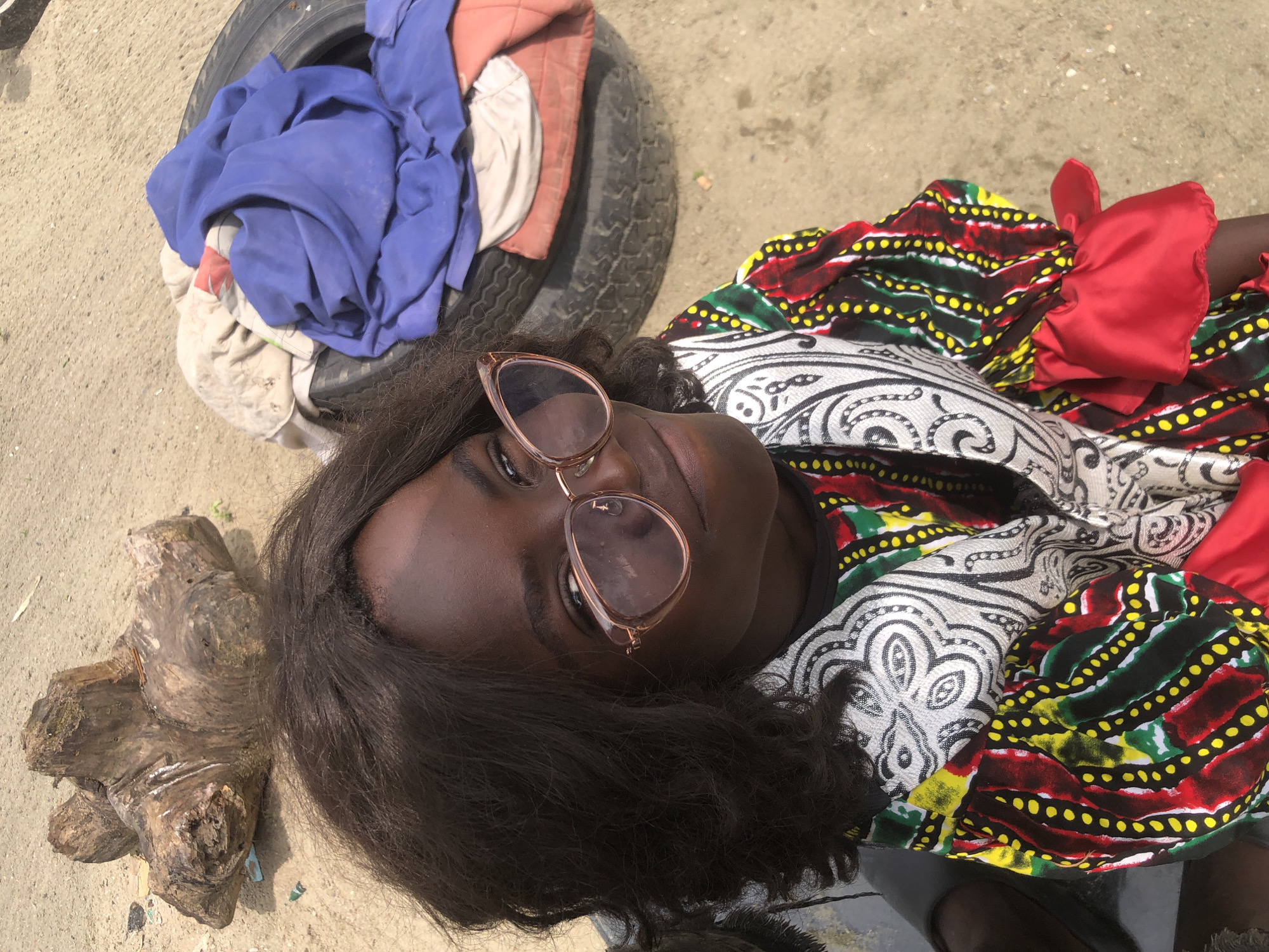The image shows a young Black female wearing glasses and a colorful patterned dress. She is outdoors, on what appears to be a sandy or dirt surface. In the background, there is a weathered piece of wood and a tire with several pieces of fabric piled on top of it. The overall impression is that of an outdoor setting in a possibly informal or rural area.