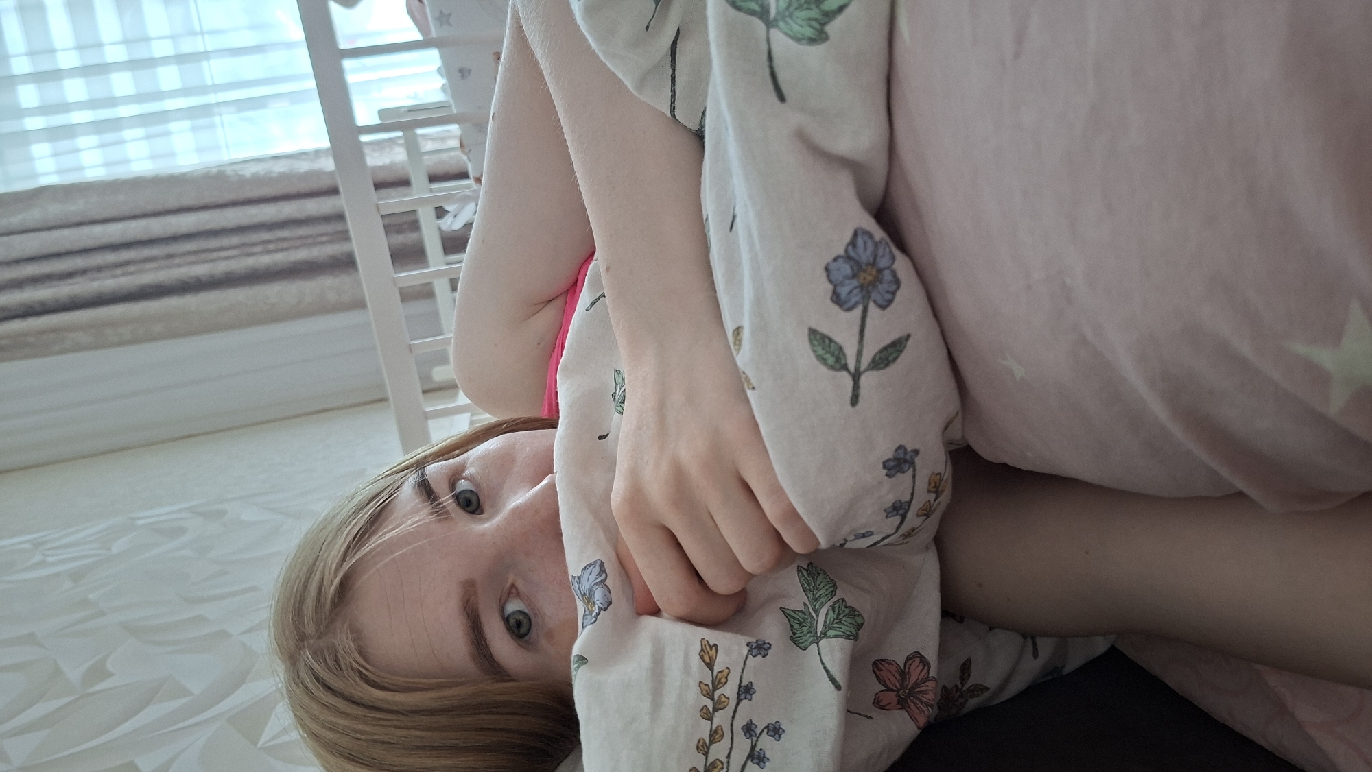 A young blonde woman with blue eyes lies in bed, holding a patterned blanket close to her face. The blanket is predominantly white with floral prints in various colors, including blue, green, and orange. A pink pillow or blanket is also visible. The woman is lying on a white, textured surface, possibly a bedsheet. In the background, a white crib-like structure is partially visible, with light coming through a window. The location is Chaikovsky, Russia.