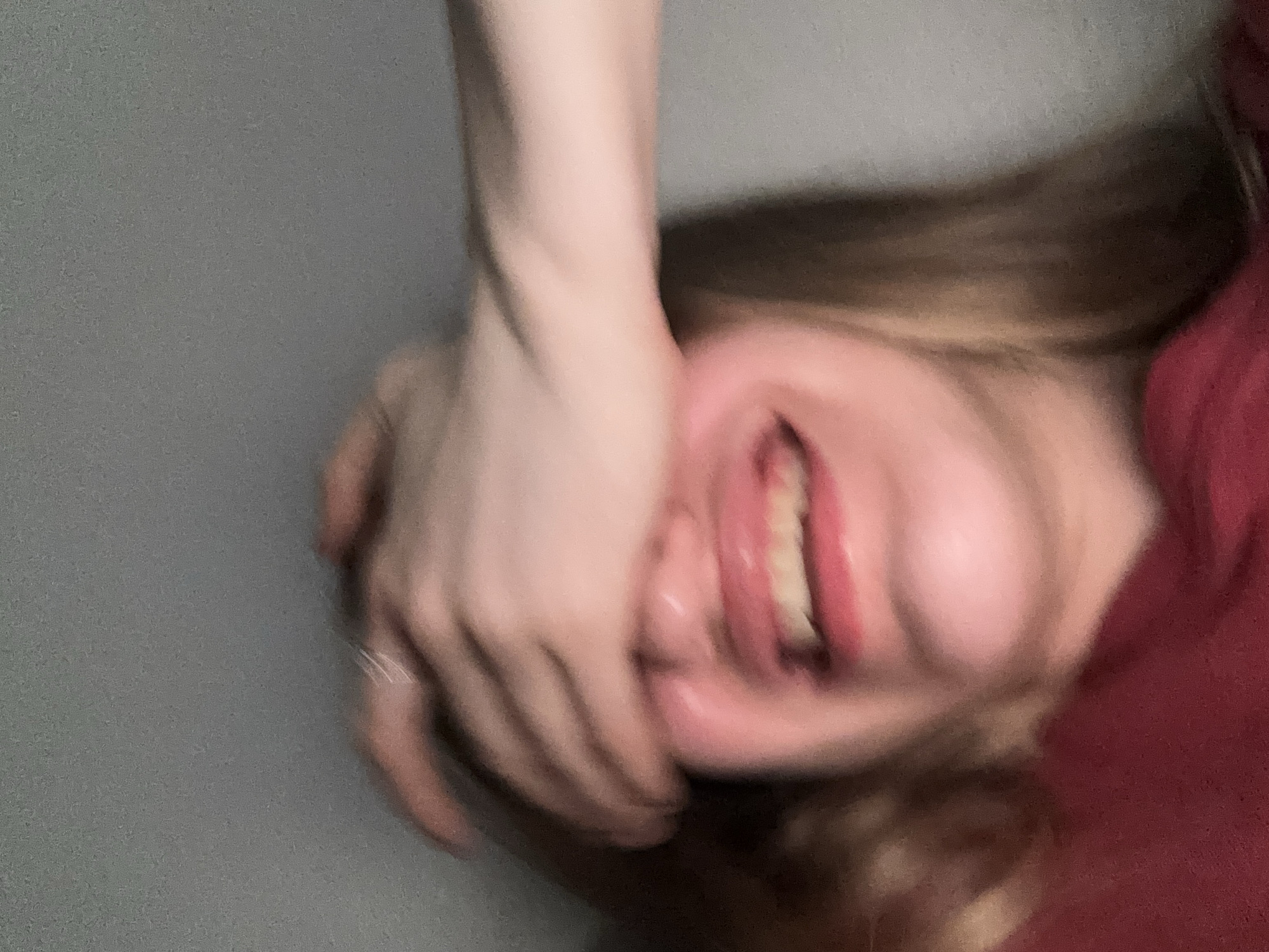A close-up, blurred image shows a person's face partially covered by their hand. Their mouth is open in a wide smile or laugh, revealing teeth. The person is wearing a dark red top. The background is a plain, light gray wall. The image does not contain any elements that would allow for a specific location in Vsevolozhsk, Russia to be identified.