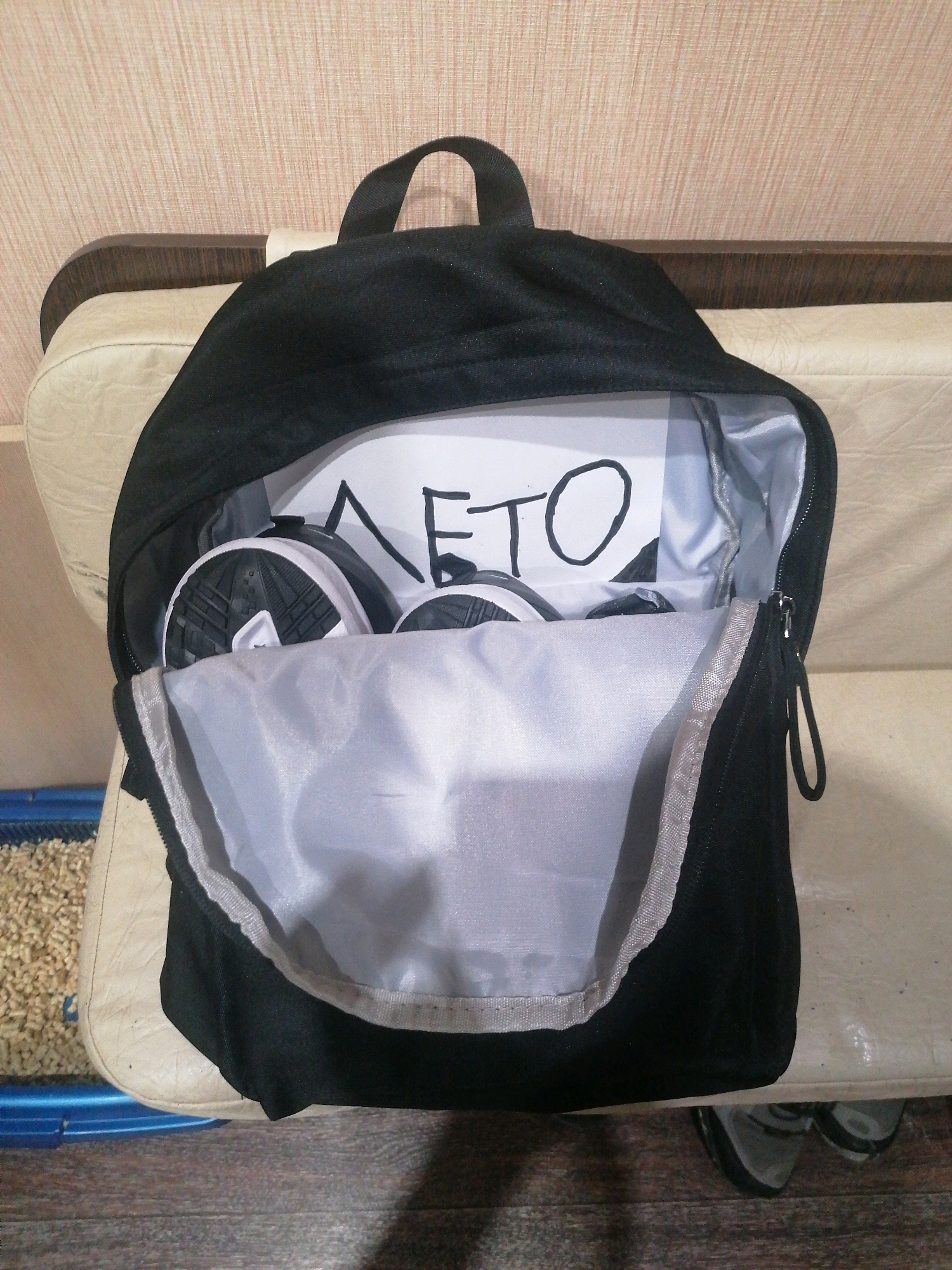A black backpack is placed on a beige sofa. Inside the backpack, a piece of paper with the handwritten Cyrillic word "ЛЕТО" (Summer) is visible. Two black and white sneakers are also inside the backpack. The backpack is partially unzipped, revealing its grey interior lining. The background shows a textured, light-colored wall and the edge of the sofa. To the left of the sofa, a blue and beige litter box filled with wood pellet litter is partially visible. The scene is likely indoors, possibly in a residential setting. There is no indication of the specific location in Angarsk, Russia.