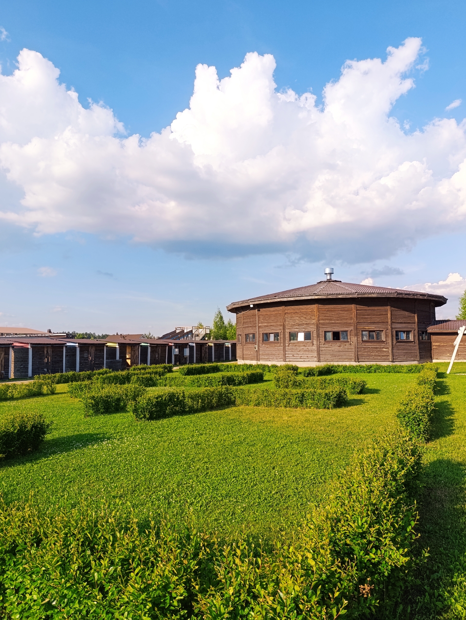 The image shows an outdoor scene in the city of Ivanovskoe, Russia. In the background, there is a large, circular, wooden building with a brown, sloped roof and several small, rectangular windows. In front of this building, there is a row of smaller, wooden structures with corrugated metal roofs, each with a different colored panel on the front. The foreground is dominated by a lush green lawn with neatly trimmed hedges arranged in rectangular patterns. The sky above is a clear blue with fluffy white clouds. The scene appears to be a recreational or park area.