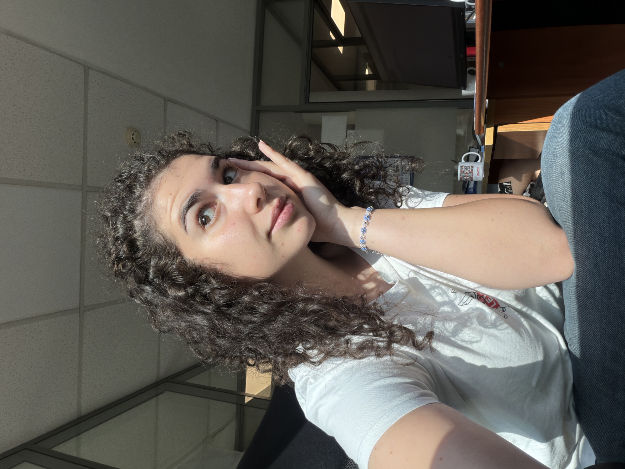 A young woman with curly brown hair is sitting in an office. She is wearing a white t-shirt and jeans. Her right hand is resting on her cheek, and she is looking to her left. Sunlight is streaming in from the right side of the image, casting shadows on her face and t-shirt. In the background, there is a desk with a computer monitor, a coffee mug, and some papers. The office appears to be in a modern building with glass partitions. There is a smoke detector on the ceiling. The overall impression is that of a casual, everyday moment captured in an office environment. The location is not specified in the image.