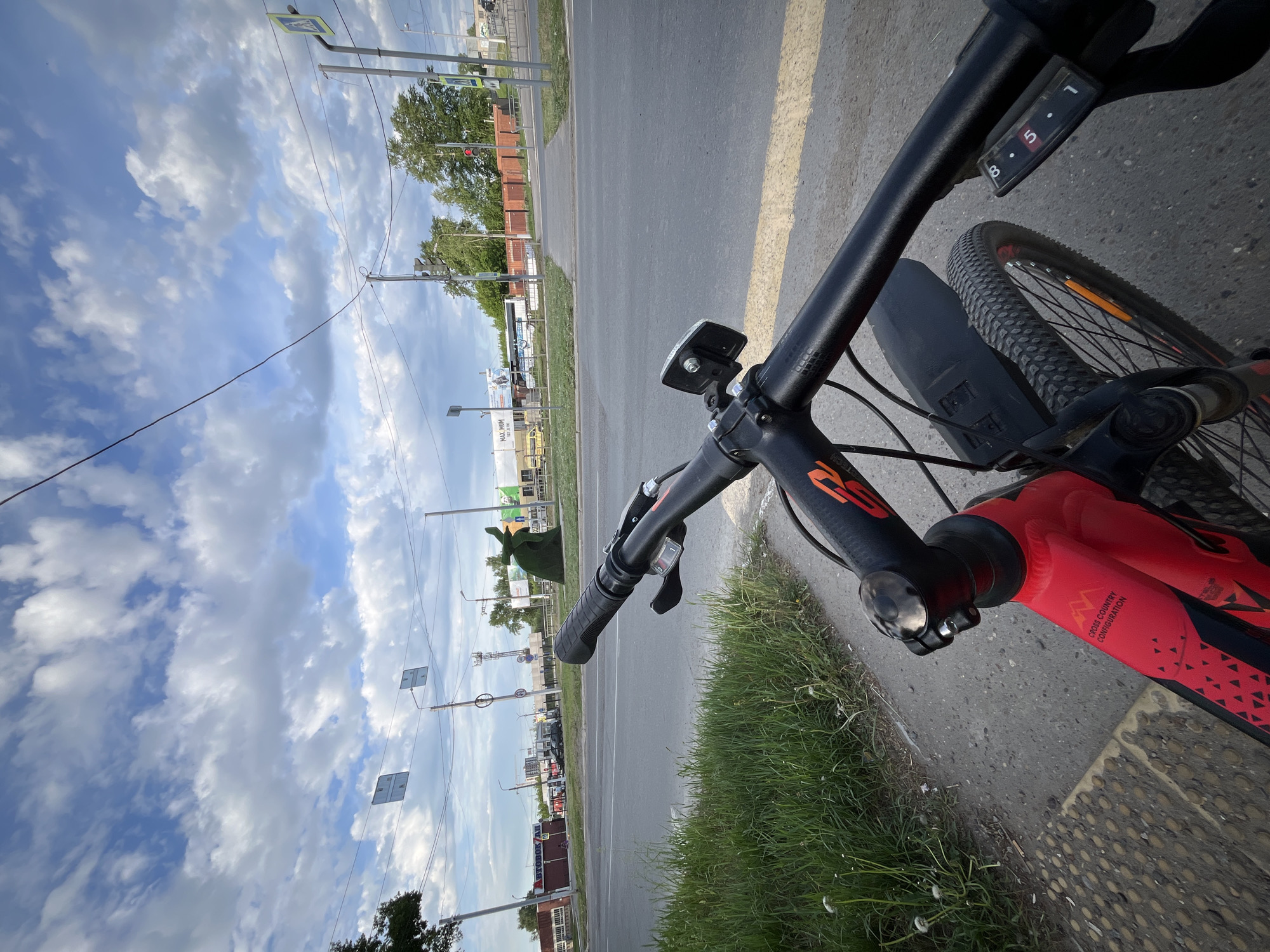 View from the handlebar of a red mountain bike, possibly near the intersection of Sovietskaya street and Voskresenskaya street in Chaikovsky, Russia. The bike is on a sidewalk, with a strip of grass between the sidewalk and a yellow-striped road. Buildings are visible across the road. The sky is partly cloudy.