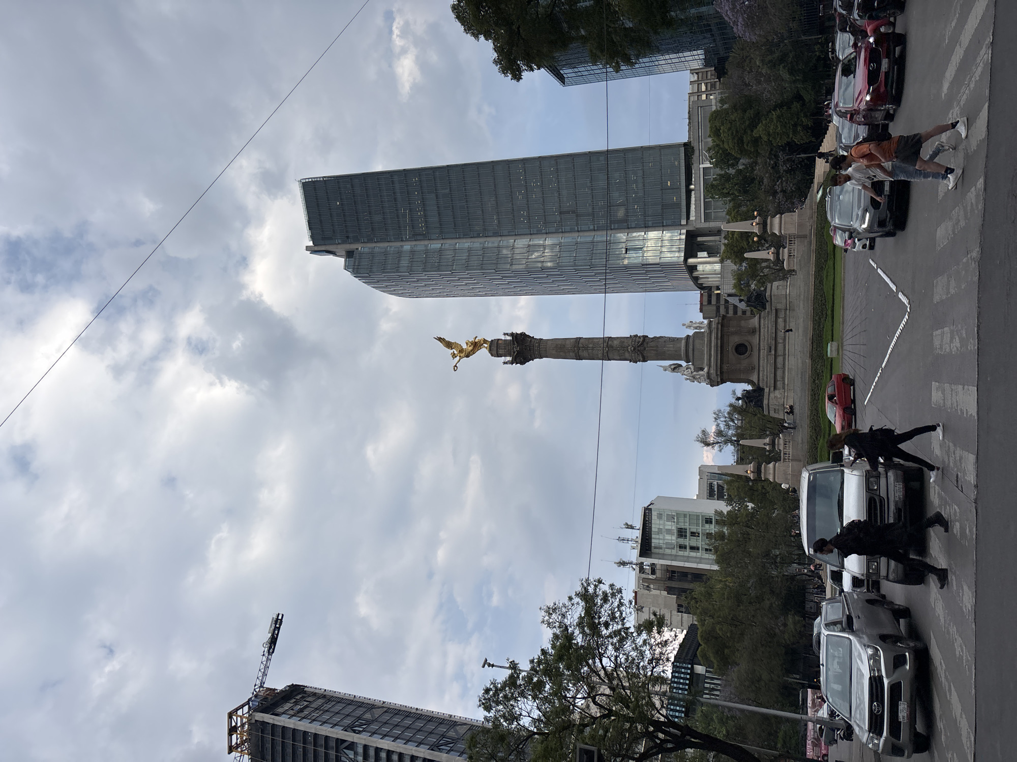 This image captures a vibrant urban street scene in Mexico City, centered around the iconic Angel of Independence monument. The scene is set during the daytime under an overcast sky with significant cloud cover.
In the mid-ground, the gilded statue of the Winged Victory, known as the Angel of Independence, stands atop its tall column, serving as a prominent landmark. It is situated on a large traffic circle or glorieta, surrounded by manicured green spaces and decorative balustrades.
In the foreground and to the right, several individuals are actively crossing a wide street on a marked pedestrian crosswalk. They are dressed in casual attire. Numerous vehicles, including pickup trucks and cars, are visible on the street, either parked or in motion, indicating typical urban traffic.
The background is dominated by modern architecture. A tall, glass-clad skyscraper rises prominently behind the Angel of Independence. To the left, another tall building is under construction, with a large construction crane visible at its apex. Other multi-story buildings and lush green trees are interspersed throughout the urban landscape. A thin utility wire or power line spans diagonally across the sky in the upper left portion of the image.
Visible text includes partial signage on a white building in the mid-ground, which appears to read "CITY EXPRESS" and possibly "LA PAZ PLAZA". The overall atmosphere is that of a busy, yet ordinary, day in a major metropolitan area.