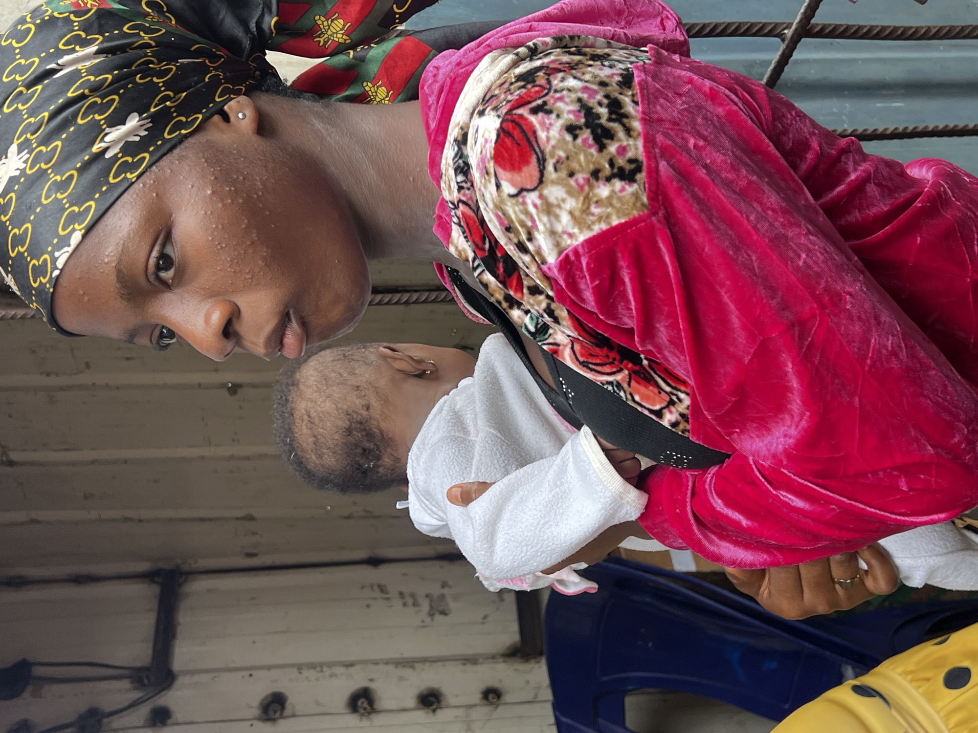 In Nkpor, Nigeria, a young Black woman, wearing a black headscarf with a gold and white pattern and a vibrant pink velvet jacket adorned with a floral print, is holding a baby. The baby, draped in a white terrycloth garment, is facing away from the camera. The woman's face is turned to the right, and her expression is thoughtful, with noticeable acne on her cheeks. A small silver stud earring is visible in her left ear. Her right hand, adorned with a gold ring, gently supports the baby. The background consists of a corrugated metal wall, suggesting an outdoor or semi-enclosed urban environment. To the left, a blue plastic chair is partially visible. The overall scene captures a moment of quiet intimacy between the woman and the baby amidst the hustle of the city.