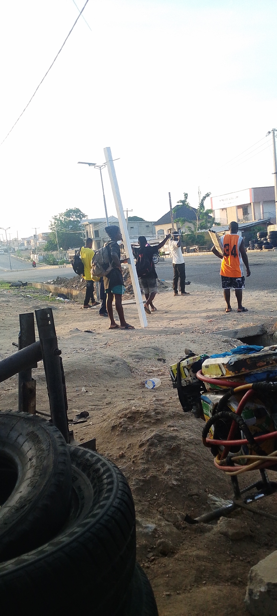 The image depicts a scene in Lokoja, Nigeria, likely near a roadside mechanic's area. A group of men are standing in the road. One is holding a long, white rectangular object. Others have their hands up, seemingly gesturing. To the front of the image are tires, a metal stand, and a piece of equipment that appears to be part of a generator. The ground is dirt. In the background, there are buildings, cars, and a street light.