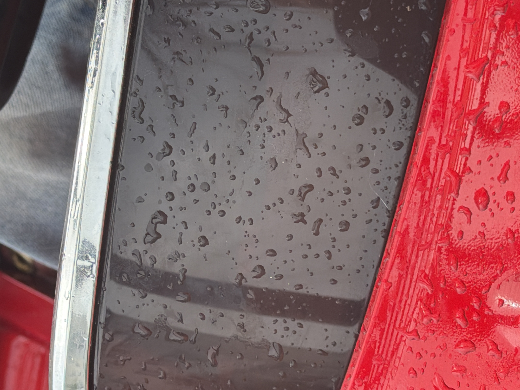 The image shows a close-up view of a car's exterior in Chaikovsky, Russia, during or after rainfall. The scene focuses on a section of the car, likely a door or body panel, which is a dark gray color and covered in numerous water droplets of varying sizes. A thin, reflective chrome trim runs horizontally across the top of the dark gray surface. Below the dark gray section is a bright red surface, also wet with smaller water droplets. The texture of both the gray and red surfaces appears smooth, but the water droplets create a speckled, uneven appearance. The upper portion of the image shows a hint of blue denim fabric, suggesting a person is in the vicinity, possibly sitting in the car or leaning against it. The lighting is diffused, indicating an overcast or rainy day. The overall impression is one of a wet, everyday outdoor environment, possibly a street or parking area in Chaikovsky.