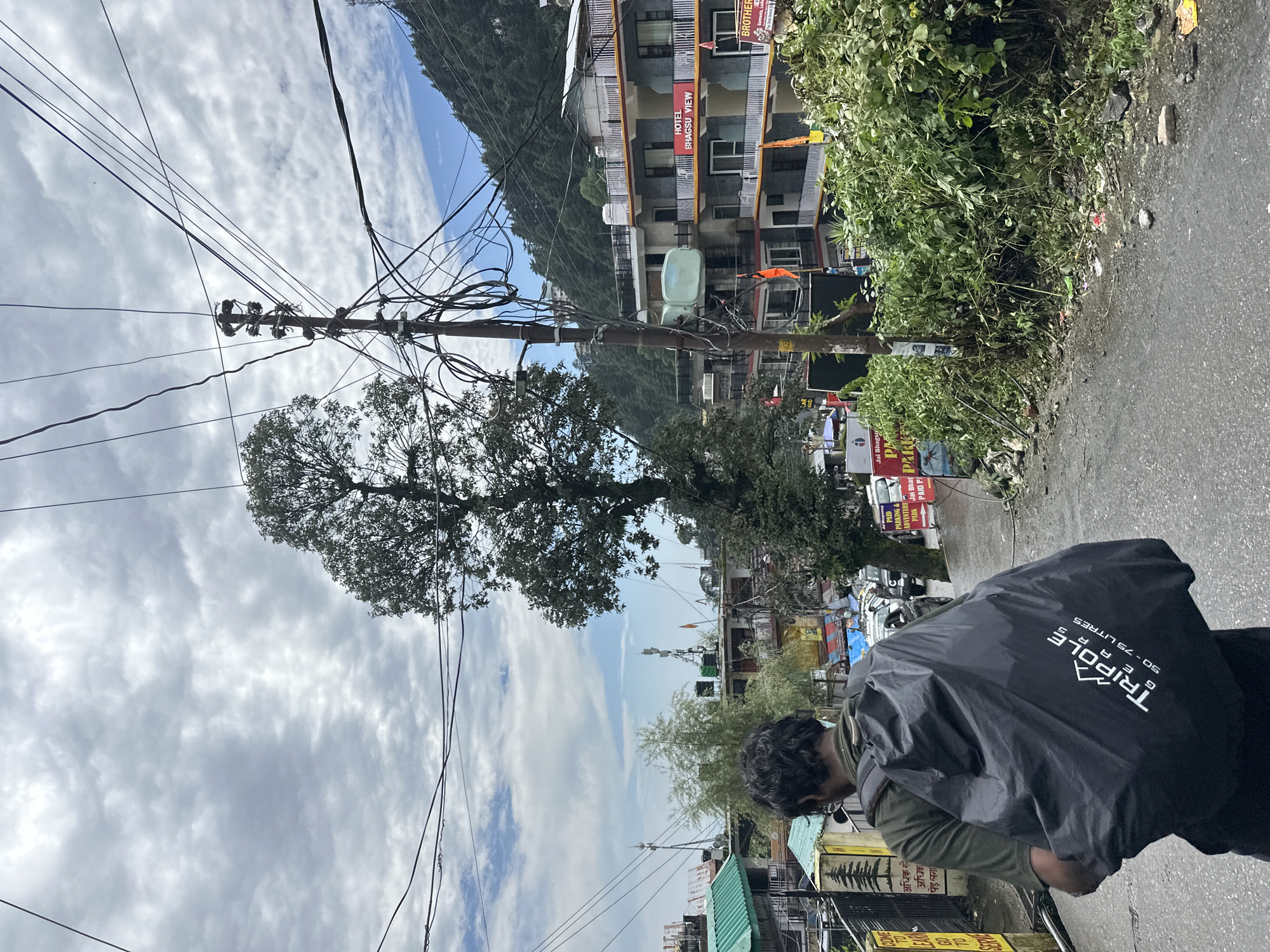 The image captures a street scene in a hilly urban area, likely India given the signage. A person wearing a green shirt and glasses walks away from the camera, carrying a large black backpack with "Tripole Gears 50-75 Litres" printed on it. The backpack is covered by a black rain cover. The street is paved and wet, suggesting recent rain.
On the right side of the street, a multi-story building labeled "HOTEL BHAGSU VIEW" is visible. The building has several balconies with white railings and some have red banners. Another banner indicates "BROTHERS". Orange flags are also present on the building.
To the left of the street, there are smaller buildings with green roofs and some yellow signage. A large tree stands prominently in the middle ground. Overhead, a complex network of electrical wires crisscrosses the sky, attached to a utility pole. In the background, a steep, tree-covered mountain slopes upwards. The sky is cloudy with patches of blue. Debris is scattered along the side of the road, particularly near a dense cluster of green bushes on the right. Several cars are parked or driving on the street.