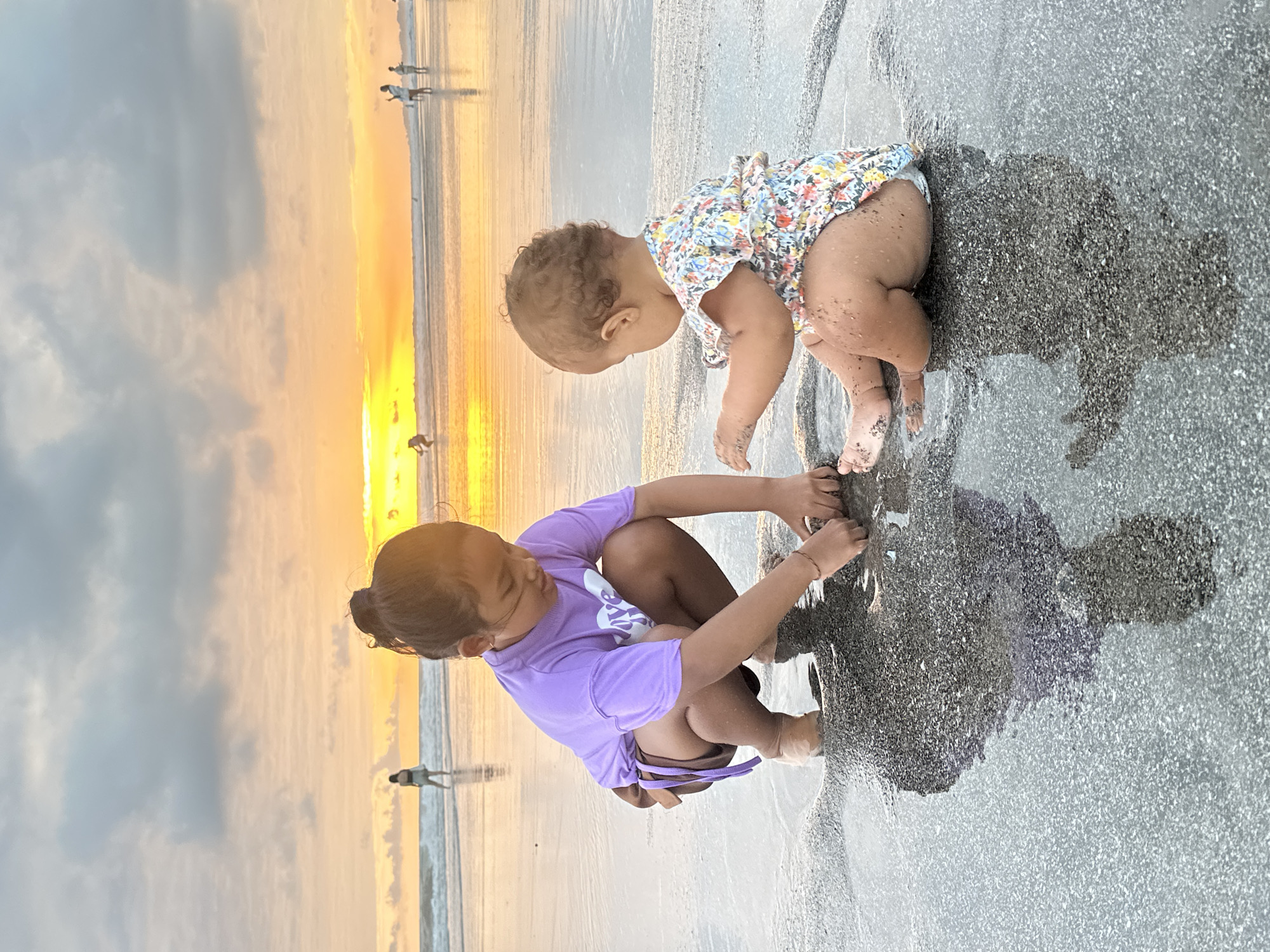 Two young girls are playing in shallow water on a beach in North Kuta, Indonesia, during a sunset. The older girl, wearing a purple t-shirt and brown shorts, is crouched down and digging in the wet sand. The younger girl, wearing a floral-patterned romper, is sitting in the water to her right, also with her hands in the sand. The sky is filled with dramatic clouds illuminated by the setting sun, casting a warm glow over the scene and reflecting on the wet sand. In the background, several other people can be seen walking along the shoreline and in the water. The tide is out, leaving a wide expanse of wet sand.