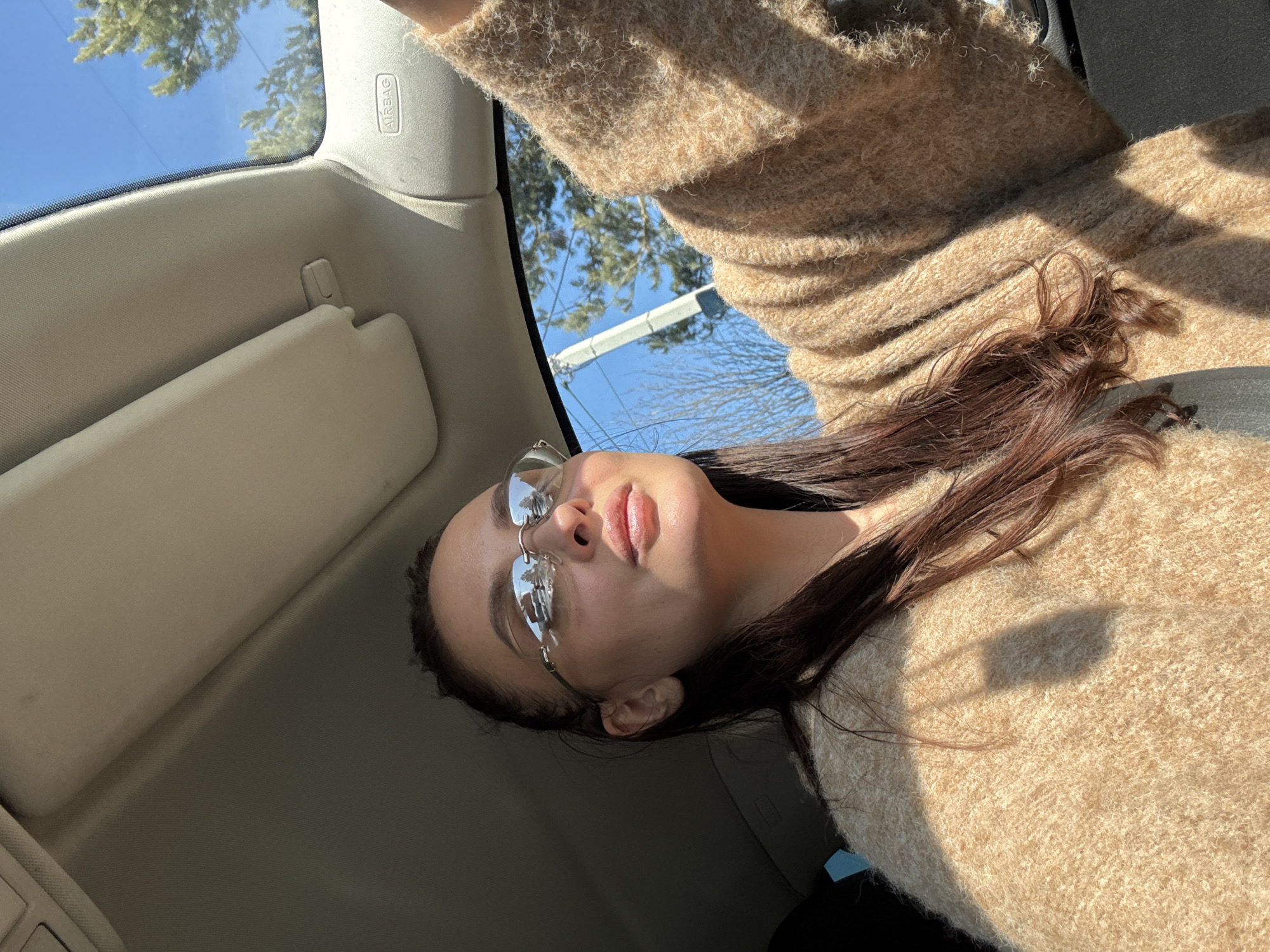 A young woman with long brown hair, wearing rimless glasses and a beige sweater, is seated in a car. Sunlight illuminates her face. The car's interior is visible, including the airbag label and sun visor. A glimpse of the sky and some trees is visible through the windshield. The specific location within the city of Chaikovsky, Russia, cannot be determined from the image.
