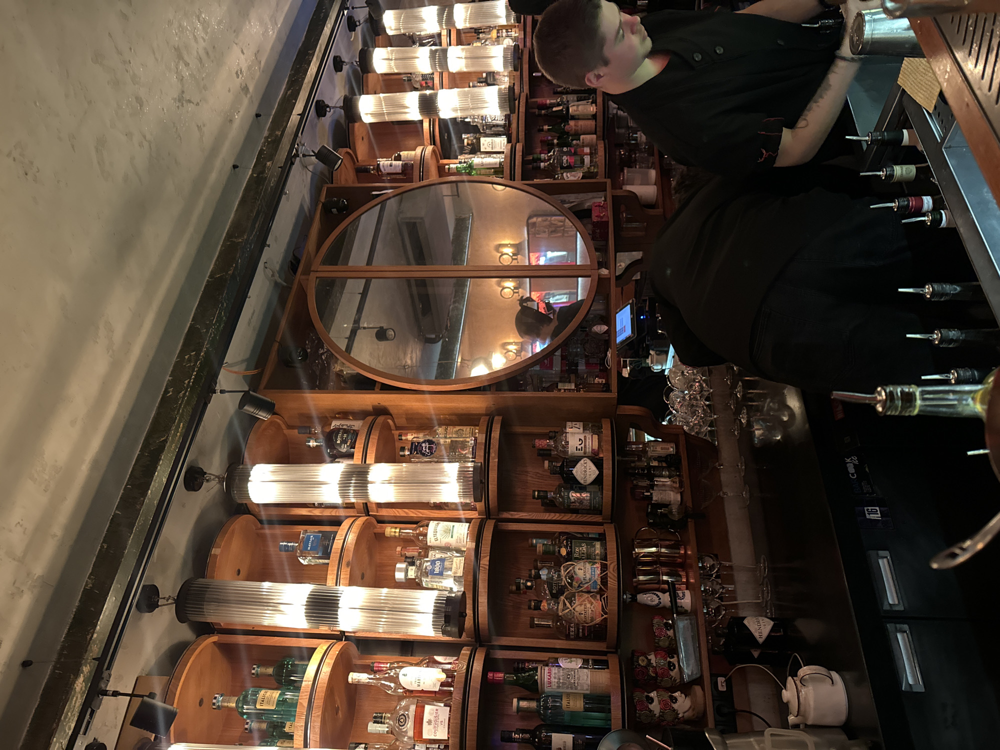 The image displays the interior of a bar located in Moscow, Russia, focusing on the back bar and the counter area. The scene is illuminated by multiple overhead cylindrical fluted glass pendant lights and track spotlights, casting a warm glow on the wooden shelving. The back bar features a modular arrangement of curved, light-colored wooden shelves densely populated with hundreds of liquor bottles of various brands, including gins, tequilas, whiskeys, and rums. A prominent, large circular mirror with a wooden frame is centrally positioned on the back wall, reflecting a dimly lit section of the bar with additional lighting fixtures and a blurred human figure.
Two individuals are present behind a dark-colored bar counter. On the right, a young male with short, light brown hair, wearing a black chef-style jacket with a red emblem on the sleeve, is leaning over the counter, looking downwards, actively engaged in a task involving bar tools or drink preparation. To his left, partially obscured, another individual in dark clothing is bent over the counter, suggesting interaction with equipment or items below the counter level.
The bar counter surface displays various tools and items including small bottles with pour spouts, a metal cocktail shaker, glassware, and a small white teapot. Below the counter, dark refrigeration units are visible, one marked "BAR COOK 100". On lower shelves behind the counter, decorative items such as two stylized ceramic head busts with floral patterns, copper barware, and additional bottles and glassware are arranged. The ceiling above is a textured, light-colored surface crossed by a dark metal beam and a lighting track.