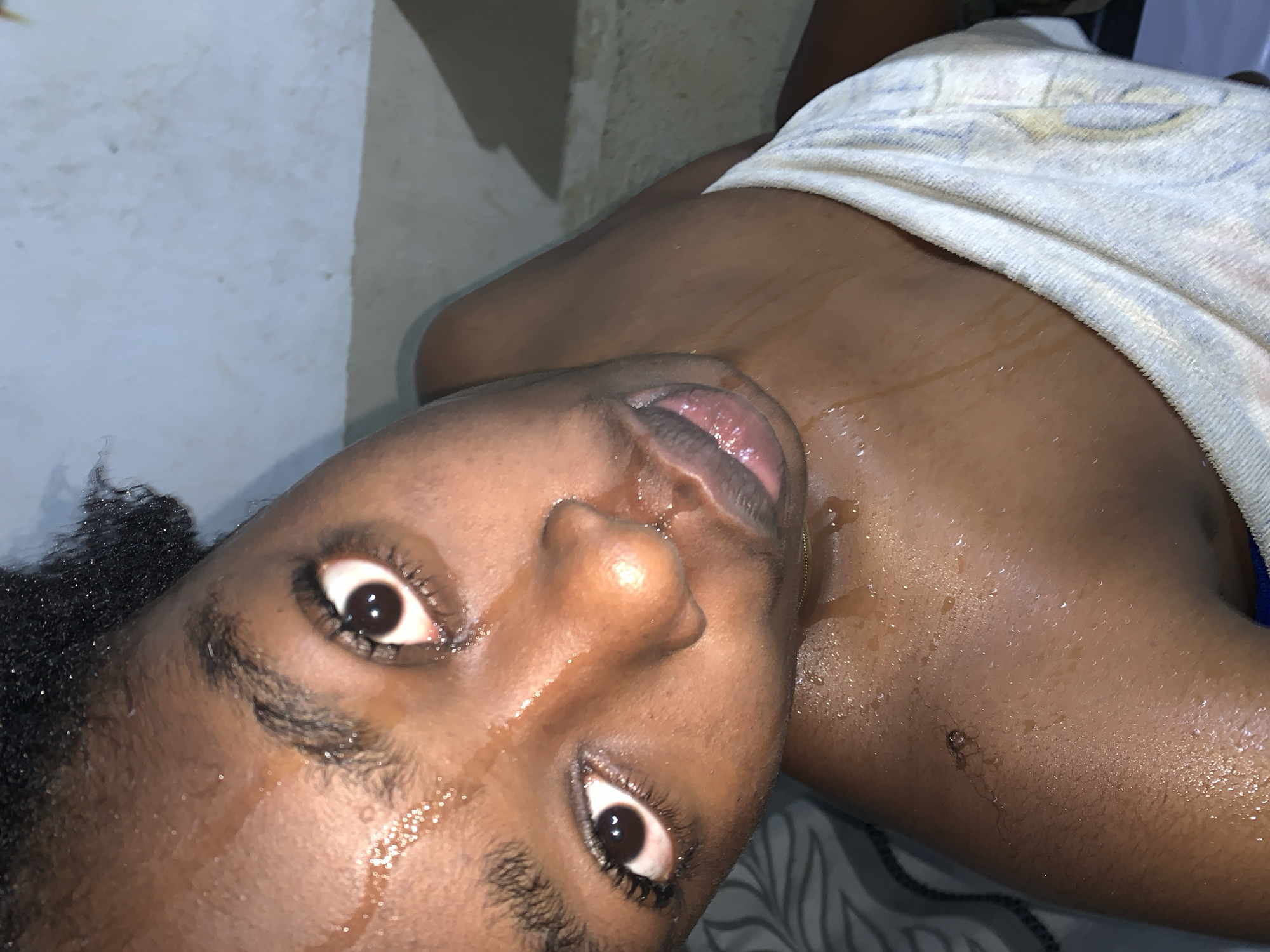 The image shows a close-up, overhead view of a Black person's face and upper torso. The person appears to be young, with dark skin that is glistening with sweat or water. Their hair is dark and textured, pulled back from the face. The person's eyes are open and looking upwards. Their mouth is slightly open, with the tip of their tongue visible. They are wearing a light-colored towel or cloth draped around their shoulders. The background appears to be a wall, possibly in a bathroom or similar setting, with some discoloration and marks. The lighting is somewhat dim, creating shadows and highlighting the wetness on the skin. The image is taken from a high angle, looking down at the subject. There is no specific information about the location in Rafin Par, Nigeria, that can be inferred from the image itself.