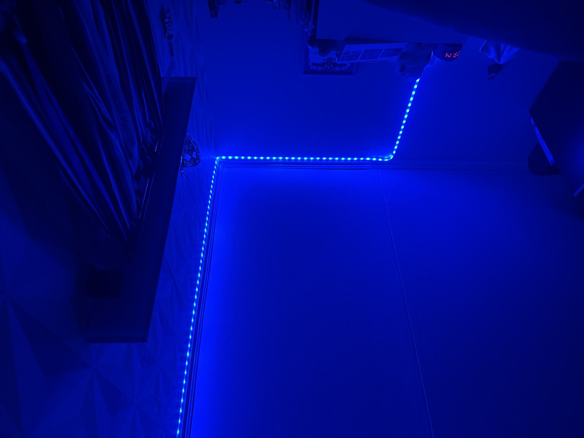 The image shows a room illuminated by blue LED strip lights. The lights run along the edges of the walls and ceiling. There is a wardrobe or closet on the left side of the image. There are curtains on the left side, and a geometric patterned wall. On the top right is an air conditioner and an alarm clock. The room appears to be a bedroom. The image was taken in Chaikovsky, Russia.