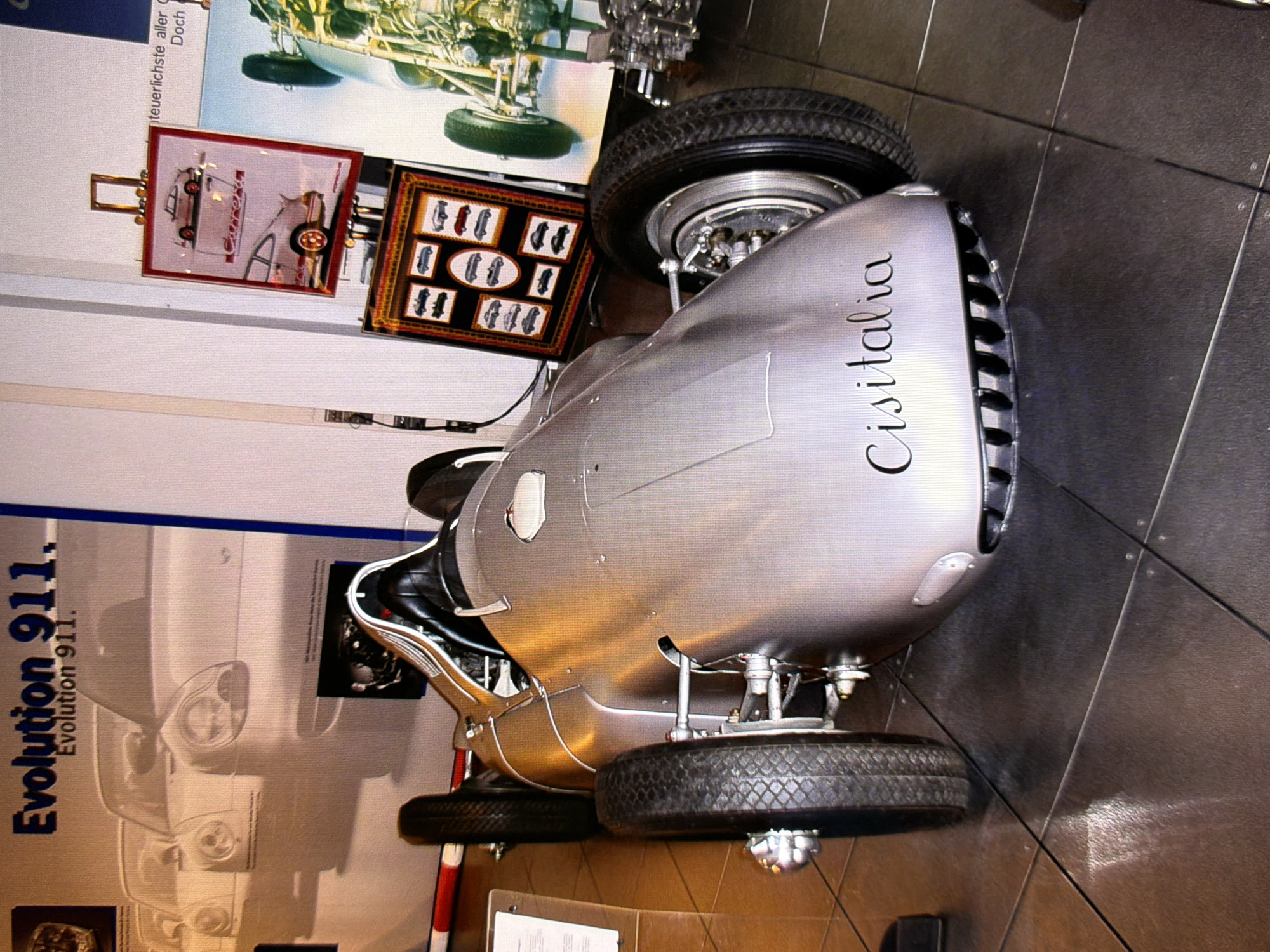 The image shows a silver Cisitalia race car parked indoors on a dark tiled floor. The car has a sleek, aerodynamic design with a single seat and exposed mechanical components. Behind the car, a poster displays images of Porsche cars with the text "Evolution 911." Another display to the right shows a framed collage of smaller car images. The environment appears to be a museum or exhibition hall. There are no people visible in the image. The location is identified as Kyiv, Ukraine.