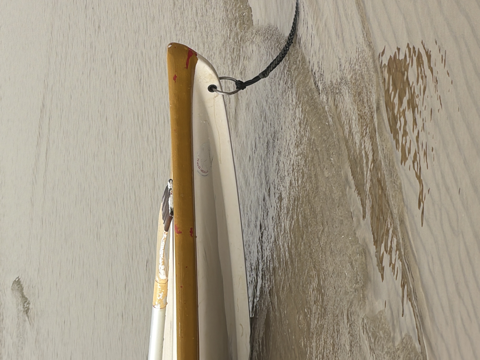 This vertical close-up image features the side of a small boat, likely a canoe or kayak, positioned in or alongside shallow water near a textured, wet sandy or muddy surface. The boat is predominantly white or off-white, with a prominent wooden-colored trim along its gunwale, which displays some reddish marks, possibly from wear or paint. A metal ring is attached to the boat, through which a black braided rope is secured with a knot, suggesting it is moored or tied.
On the left, a white-shafted oar or paddle with a yellow grip and a black and white blade is partially visible, resting against the boat. No people are present. The surrounding surface exhibits distinct ripple or wave patterns, indicative of shallow water movement. On the right side, darker, irregular patches suggest areas of deeper saturation or standing water, adding to the textured appearance.
Faint Hebrew text is visible on the boat's hull, reading "נוח" (Noach) and "תל אביב" (Tel Aviv), confirming the boat's origin or registration in Tel-Aviv, Israel. The scene is illuminated by natural light, consistent with daytime, highlighting the textures of the water and boat surfaces. The overall impression is one of quiet stillness, with the boat stationary in its aquatic environment.