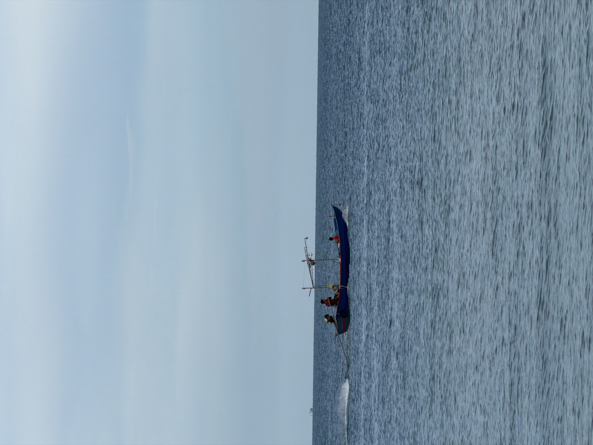 A blue and red fishing boat with a canopy is traveling through calm, blue ocean water under a pale blue sky. Three individuals are visible on the boat, two seated and one standing. One person is at the stern, another in the middle of the boat wearing a red shirt, and a third person is at the front wearing a red shirt. The boat is creating a white wake behind it. In the distance, a faint outline of land or another boat is visible on the horizon. The scene appears to be in the coastal waters of Baan Chaweng, Thailand.