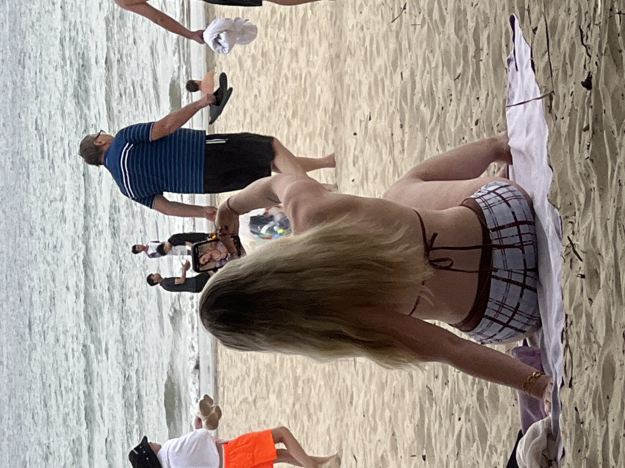 The image shows a crowded beach in Da Nang, Vietnam. In the foreground, a blonde woman in a bikini and plaid shorts is sitting on a towel and taking a selfie with her phone. Behind her, several people are walking along the shore. A man in a striped shirt and shorts is holding a pair of flip-flops. In the background, people are swimming in the ocean, and a few are standing on the wet sand. The beach is covered in light-colored sand, and the water is a muted gray-blue, with small waves. The sky is overcast.