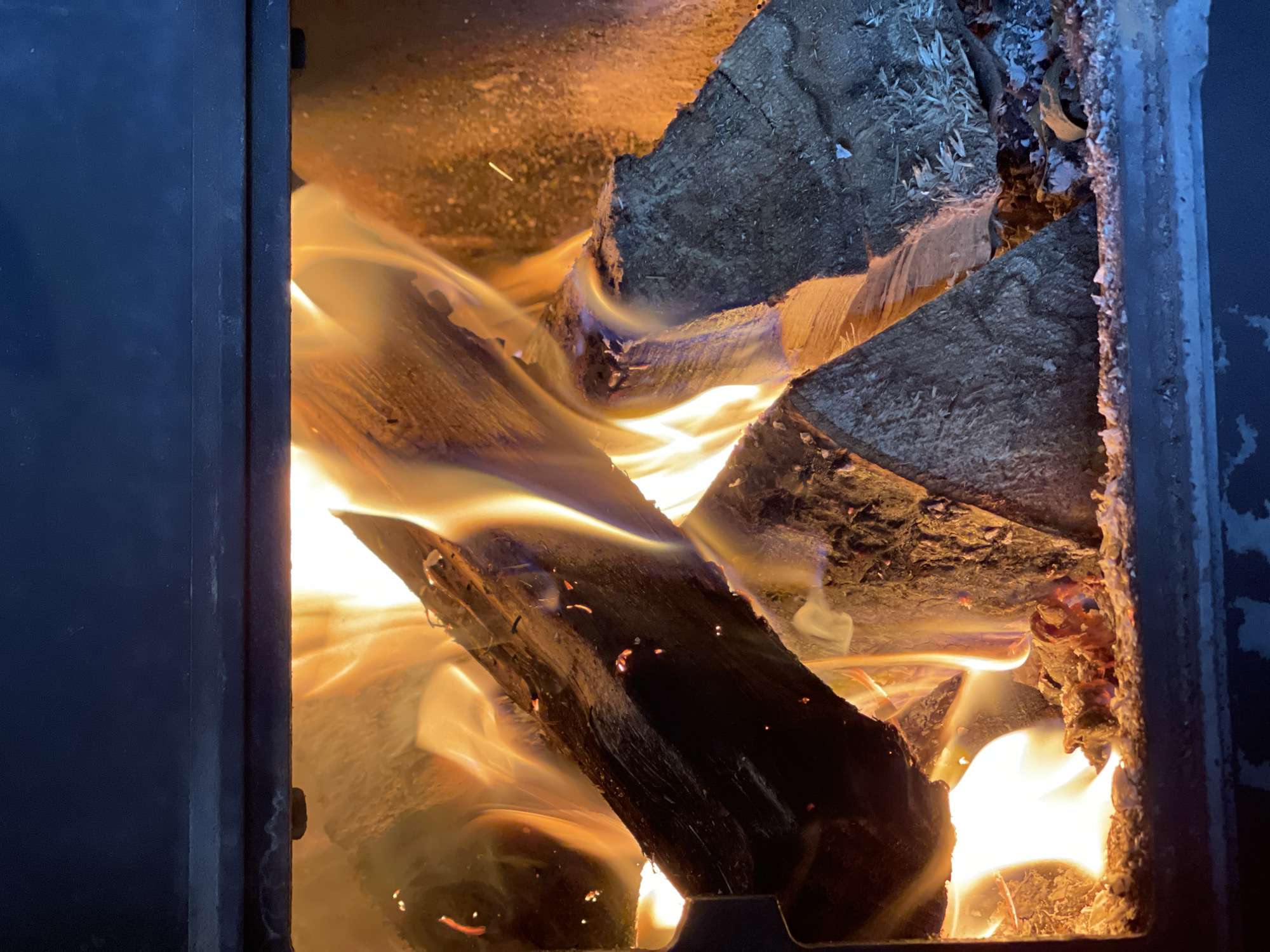 The image depicts burning firewood inside a wood-burning stove. Two large logs are visibly on fire, with flames licking around them. The inside of the stove is lined with refractory material. The stove door frame is visible on the left and right sides of the image, and the edges are black. There is no visible indication of the city of Перевозное, Russia in the image. No people are visible.