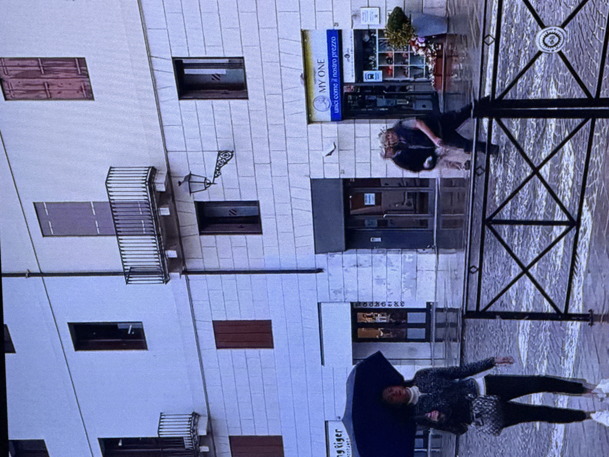 The image depicts a street scene, possibly during or after rain, with wet pavement reflecting the surrounding buildings. In the foreground, a person holding a dark umbrella walks to the left. This individual is wearing a patterned jacket over a white turtleneck and dark pants, carrying a crossbody bag. To the right, behind a black metal fence, two individuals are walking away from the camera. One is wearing a dark vest over a light shirt and dark pants, while the other is in a dark jacket and lighter pants.
The background features a white brick building with several windows and balconies. A sign above a storefront reads "MY ONE" with the tagline "unici come il nostro prezzo." Other shop entrances are visible, one of which has a sign that appears to say "NG TIGER". The scene is urban, with architectural elements suggesting a European city rather than Dubai, United Arab Emirates. There is no visual evidence to support the location being Dubai.