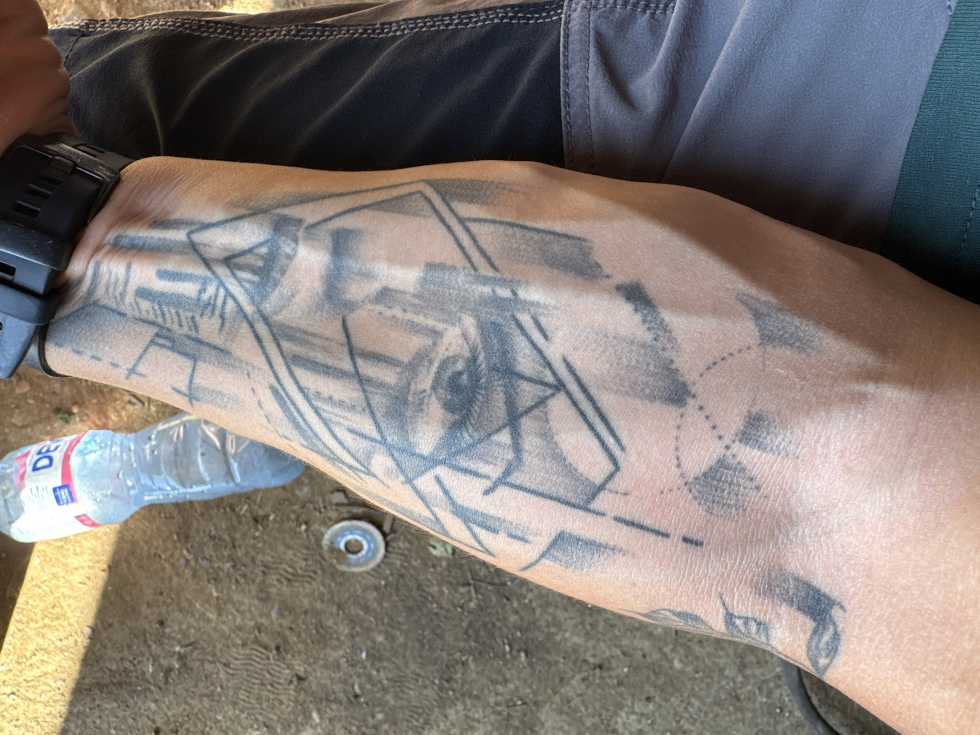 A close-up, eye-level shot captures a tattooed forearm in Prolom, Bulgaria. The tattoo, a geometric design incorporating an eye, covers a significant portion of the arm. The skin is tanned and shows visible veins. Adjacent to the forearm are grey shorts with visible stitching and a black watchband with a digital display. In the background, a partially visible plastic water bottle with a red and blue label rests on a textured, sandy ground. A small, metallic object with a circular opening lies on the ground near the bottle. The overall environment suggests an outdoor, possibly dusty or sandy, location.