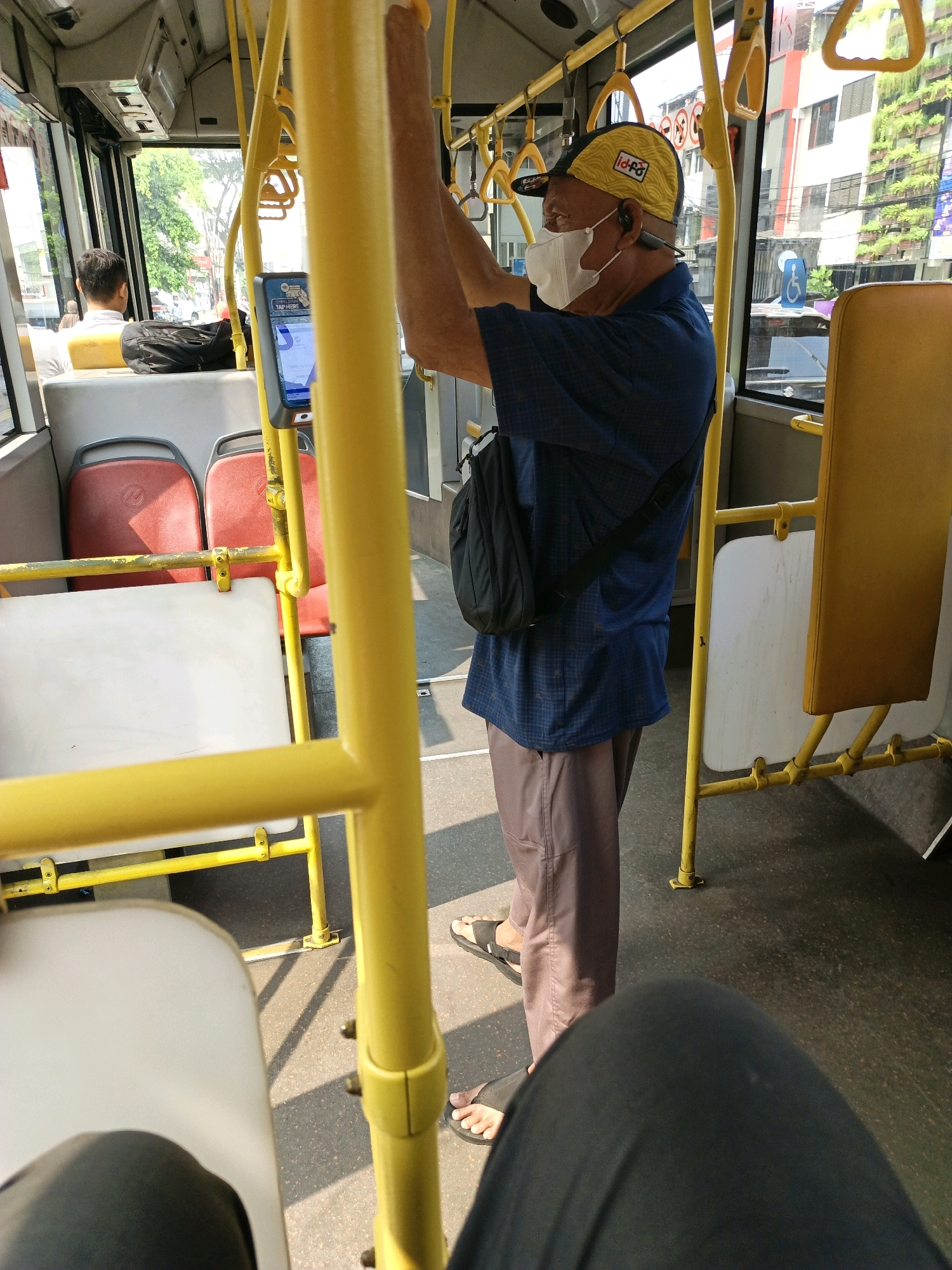 The image captures the interior of a bus in South Jakarta, Indonesia, on a daytime. A man wearing a blue checkered shirt, brown pants, a yellow cap, and a face mask stands holding onto a yellow pole. He has a black bag slung across his chest. In the background, red bus seats are visible, along with a fare payment machine mounted on a pole. Through the window, buildings and trees can be seen. The floor of the bus is dark and textured, with shadows cast by the sunlight. The perspective is from a seated passenger looking forward and slightly to the side.