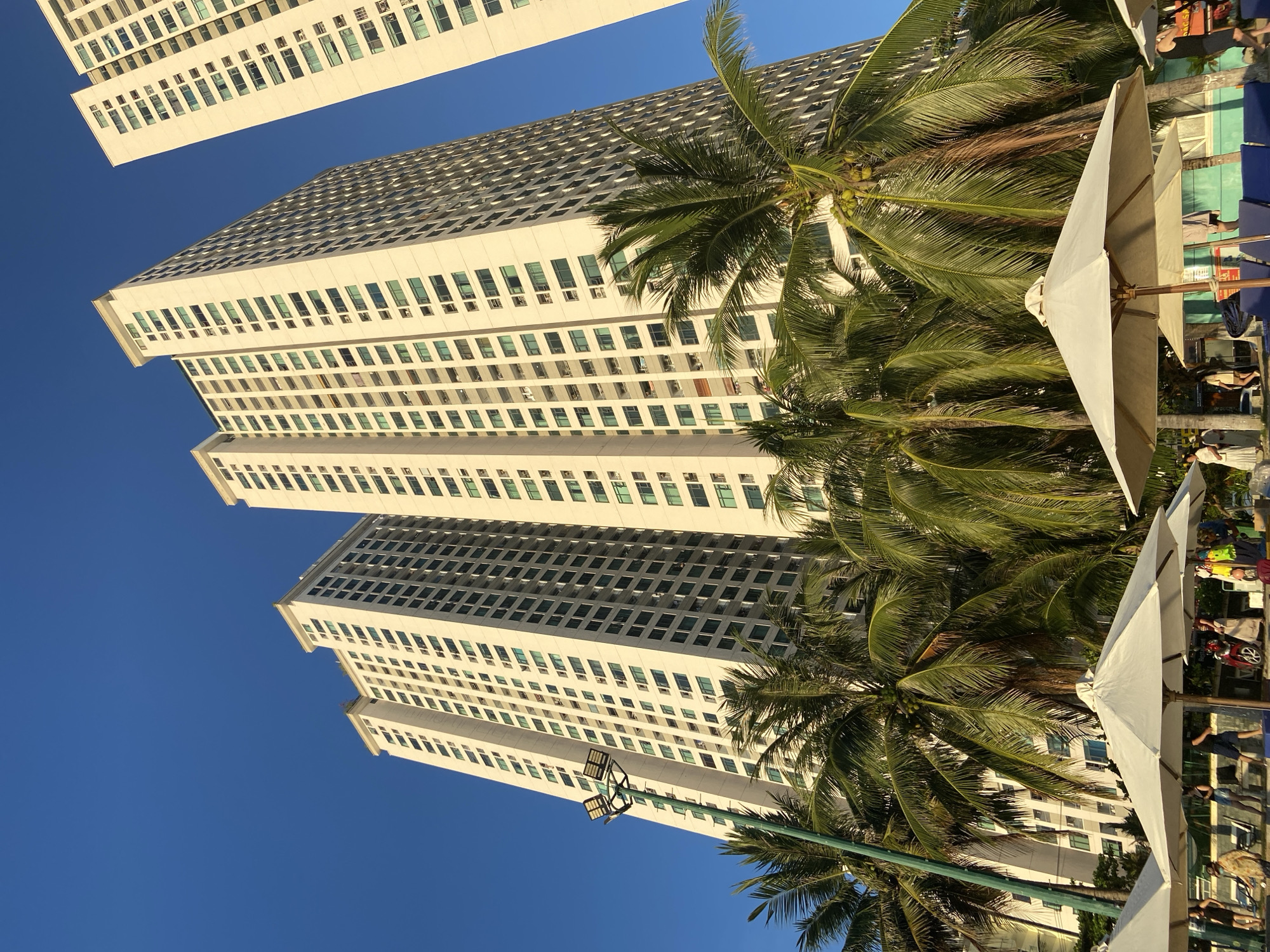 The image depicts a view of apartment buildings in Nha Trang city, Vietnam, with lush palm trees in the foreground. The buildings are tall, white, and modern, with numerous windows and balconies. The sky is a clear, deep blue. Several white beach umbrellas are set up, suggesting a resort or beachside location. People are visible near the umbrellas, some standing and others walking. The presence of palm trees and beach umbrellas implies a tropical or subtropical climate. The buildings appear to be residential or hotel complexes, characteristic of coastal tourist destinations.