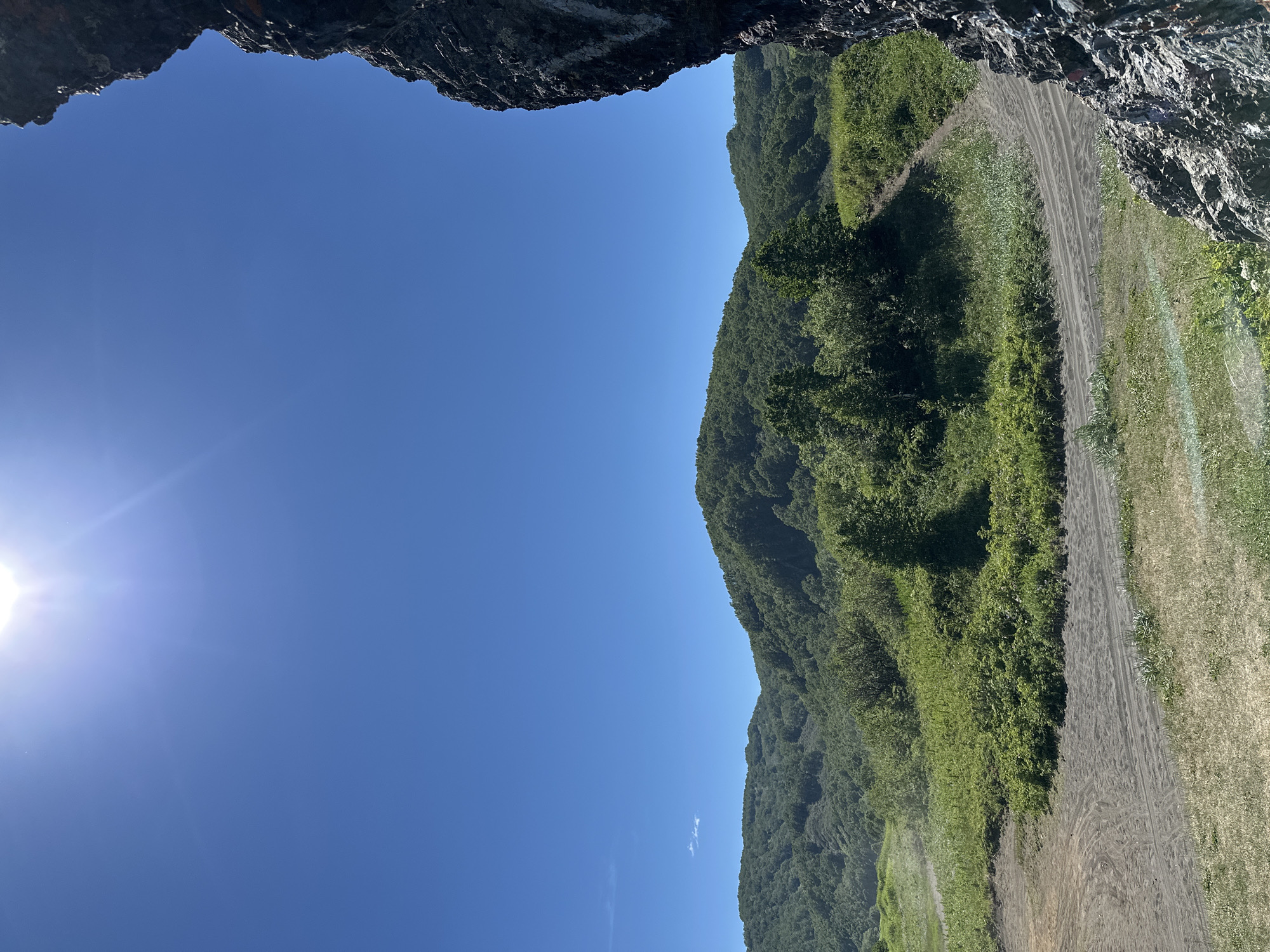 This is a landscape photograph taken on a sunny day, with a bright sun partially visible in the upper left corner of the sky. The sky is a clear, deep blue. In the foreground on the right, a dark, rugged rock face occupies the right edge of the frame, appearing as if the viewer is looking out from a cave or overhang.
The midground features rolling hills covered in dense green vegetation and trees. A dirt or sand track curves through the landscape, with visible tire tracks. The ground in this area is a mix of sand and green grass. The path leads up a gentle slope towards the wooded hills.
The background consists of more layers of green, forested hills extending towards the horizon. The overall impression is a natural, possibly remote, outdoor setting.
This scene is characteristic of the natural landscapes found in and around Petropavlovsk-Kamchatsky, Russia. The volcanic rock formations, lush green hills, and clear blue sky are common visual elements of this region. The specific location could be one of the many scenic viewpoints or trails located within the vicinity of the city, offering panoramic views of the surrounding natural beauty.