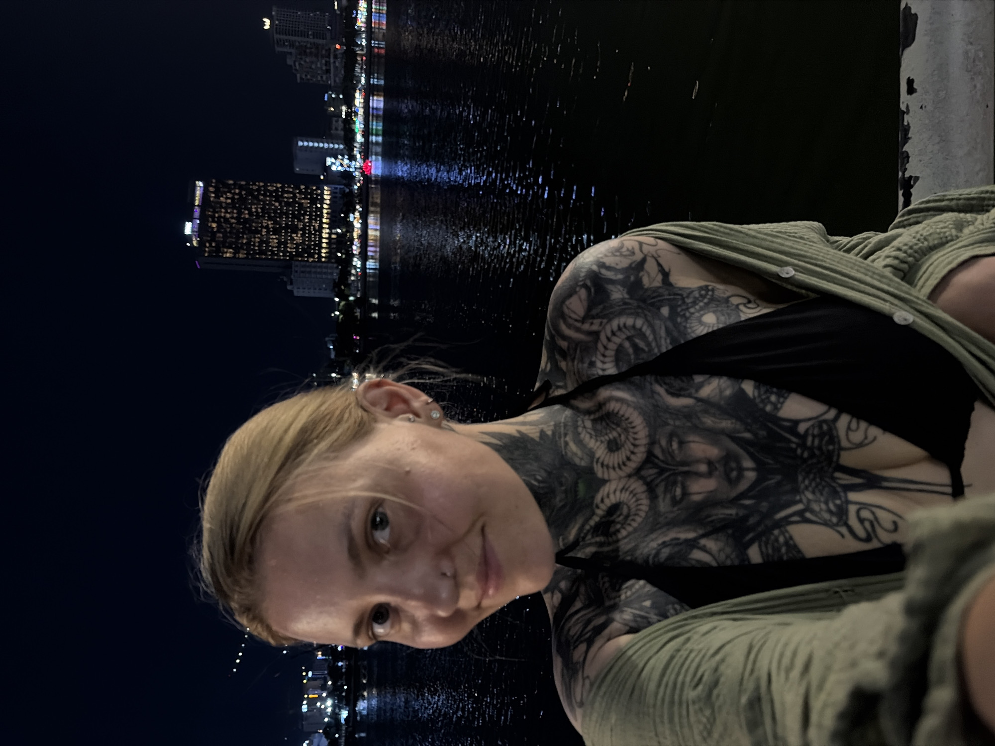 A vertically oriented selfie features a person in the foreground against a luminous night cityscape of Đà Nẵng, Vietnam. The subject, a young adult with blonde hair pulled back, looks directly at the camera with a gentle, perhaps slightly shy, smile. She has extensive blackwork tattoos covering her neck, chest, and shoulders. A prominent tattoo on her upper chest depicts an intricate design resembling a mythological head with ram-like horns and snakes. She is wearing a black top beneath a textured, olive green cardigan or shawl.
The background reveals a bustling waterfront city at night. Numerous buildings are illuminated, casting bright, elongated reflections across the dark, calm water. The scene is enveloped in darkness, indicating late evening or night. The overall impression is a casual, intimate portrait set against the vibrant urban glow of a coastal city. No text is visible in the image.