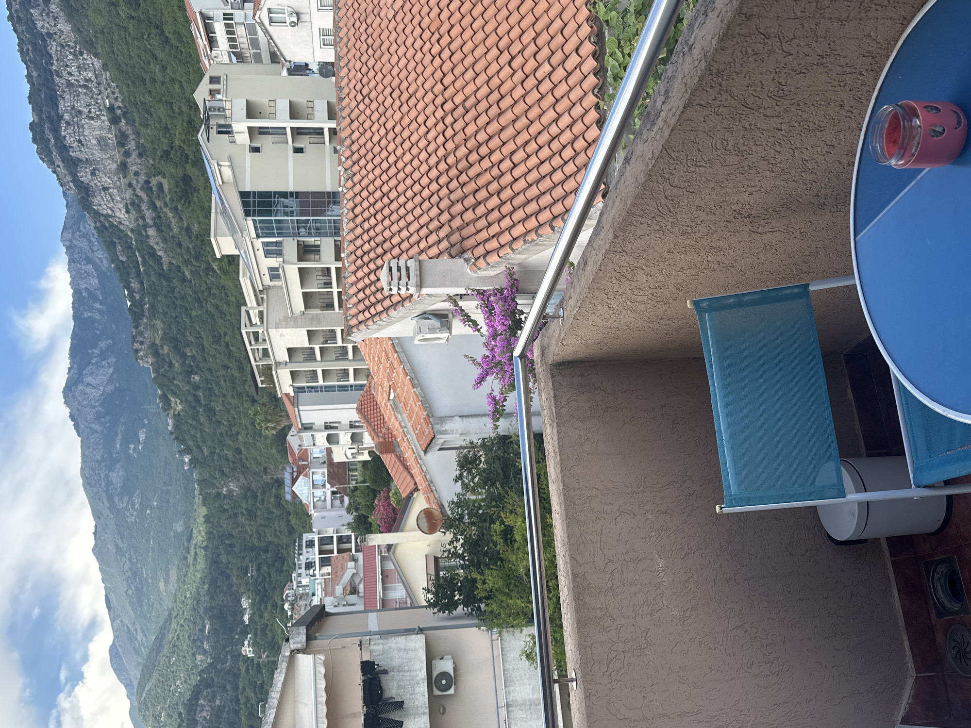 This is a media file of a view from a balcony in Budva, Montenegro, looking out over a town nestled at the base of a steep, forested mountain. The orientation of the image is rotated, with the mountain appearing on the left and the sky on the bottom. The scene is set during the daytime, under what appears to be clear weather, with scattered white clouds visible against a bright blue sky.
The foreground of the image shows a section of the balcony, featuring a textured beige wall and a metal railing. To the right, a portion of a round, bright blue table is visible, with a small, pink jar with a black lid resting on it. A folded blue fabric shade is also visible in the lower right portion of the frame.
Beyond the balcony, the view is dominated by a dense urban landscape of buildings with red-tiled roofs and white or light-colored facades. The buildings appear to be a mix of residential and possibly commercial structures, with balconies and windows visible. Further in the background, the imposing mountain rises steeply, covered in lush green vegetation. The contrast between the man-made structures and the natural landscape is striking. A cluster of vibrant purple flowers is blooming on a vine, adding a touch of color to the scene.
No people are visible in the image. The overall mood is peaceful and scenic, capturing a typical Mediterranean townscape with its characteristic architecture and surrounding natural beauty. There is no visible text in the image.