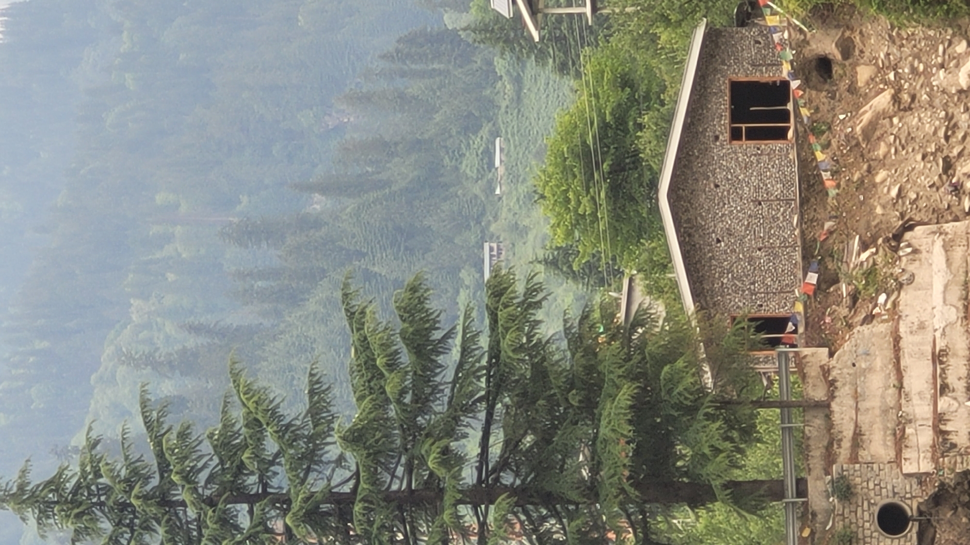 In Vashisht, India, a stone-clad building with a peaked roof sits precariously on a steep, rocky incline. The building features large, dark-framed openings that suggest windows or doorways, with some possibly serving as entrances. A string of colorful prayer flags decorates the front of the structure, fluttering in the breeze. The surrounding environment is a dense forest of evergreen trees blanketing rolling hills that disappear into a hazy, mist-covered background. In the foreground, a large evergreen tree with dense foliage stands prominently to the left of the building. Below the building, the slope is characterized by exposed dirt, rocks, and some debris. To the far left of the image, a concrete structure with a circular pipe opening is visible, integrated into the embankment. The overall scene depicts a remote, mountainous landscape with a solitary building nestled within the lush, green terrain.