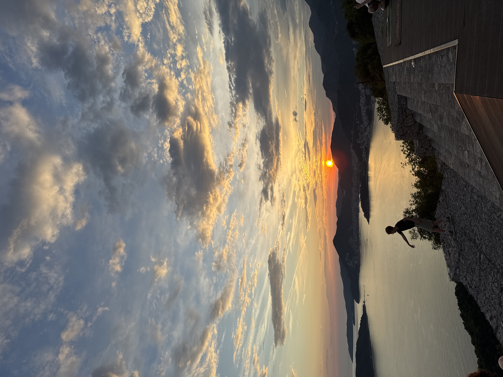 This media file captures a scenic view of a sunset over a body of water, likely a bay or sea, with mountains in the background. The sun is low on the horizon, casting a warm, golden glow across the sky and reflecting on the water. The sky is filled with dramatic clouds, some tinged with orange and pink from the setting sun, while others remain a soft blue or are darkened by shadow.
In the foreground, on a raised stone and wooden platform, a person is silhouetted against the bright sky. They are standing with their arms outstretched, seemingly taking in the view. The platform appears to be a modern structure, possibly a terrace or observation deck, with clean lines and a textured stone facade. Lush green trees are visible on the edge of the platform and along the shoreline.
The overall scene evokes a sense of peace and awe, characteristic of a beautiful sunset in a natural landscape. The location appears to be a coastal area with mountainous terrain, aligning with the context of Kamenovo, Montenegro. The time of day is clearly sunset due to the low angle of the sun and the warm, diffused light. The weather appears clear, allowing for an unobstructed view of the sunset. There is no visible text in the image.