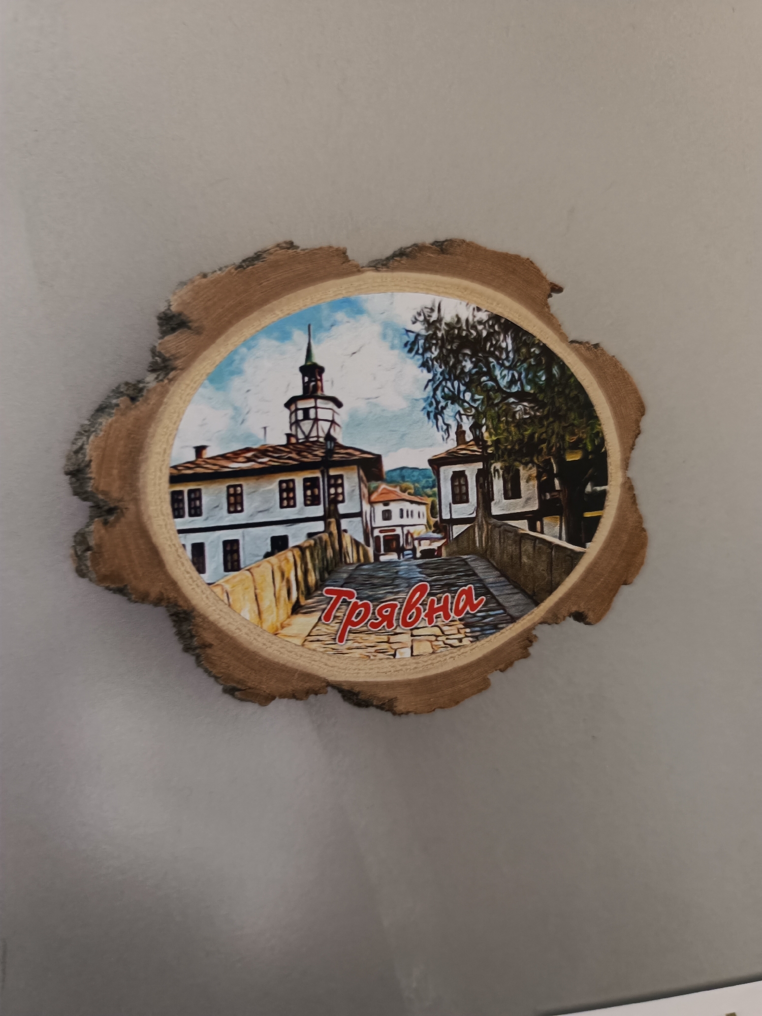 The image shows a souvenir magnet depicting a scene from Tryavna, Bulgaria. The magnet is in the shape of a rough-cut wood slice, with the edges showing bark. The scene is of a bridge and buildings in Tryavna. A building with a tall, slender clock tower is on the left. The Cyrillic word "Трявна" (Tryavna) is printed in red across the bridge. The buildings are mostly white with dark window frames. The sky is partly cloudy. The magnet is set against a neutral, grayish-beige background.